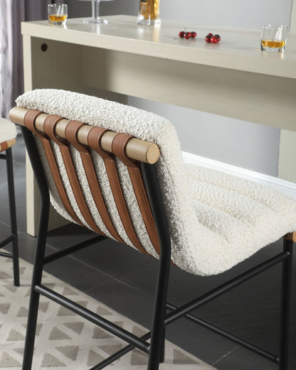The Lahni - 17" Boucle Fabric Counter Height Chair, featuring a black metal frame, light wood accents, tan leather straps, and a textured cream cushion, sits by a beige bar counter with glasses and cherries.