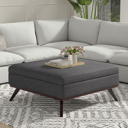 The Owen Multifunctional Square Coffee Table Storage Ottoman with wooden legs sits on a patterned rug in front of a light gray sectional, topped with a tray, books, and pink flowers beside a green potted plant in a modern living room.