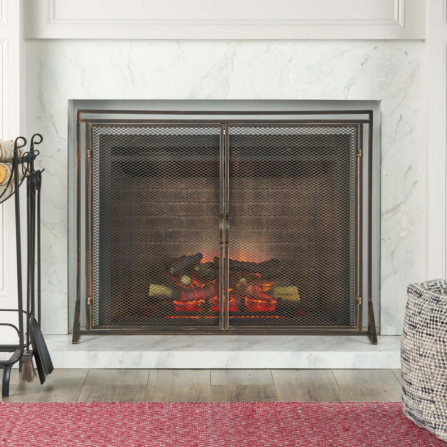 A modern fireplace features burning logs, a marble surround, and the Samantha - Iron Fireplace Screen 2 Door Design. Fireplace tools are on the left, a red rug lies in front, and a textured pouf is on the right.
