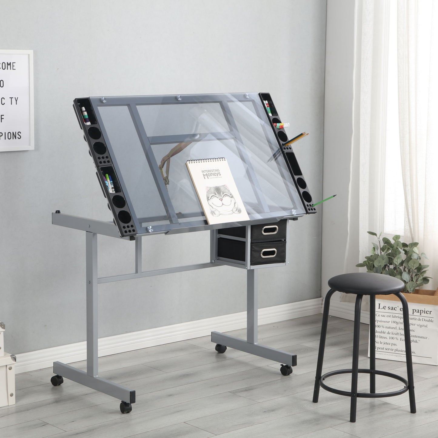The Adjustable Art Drawing Desk Craft Station Drafting With 2 Non-Woven Fabric Slide Drawers And 4 Wheels - Silver holds a sketchbook and pencils, with storage drawers beneath. A black stool sits on a light wood floor by a window with curtains.