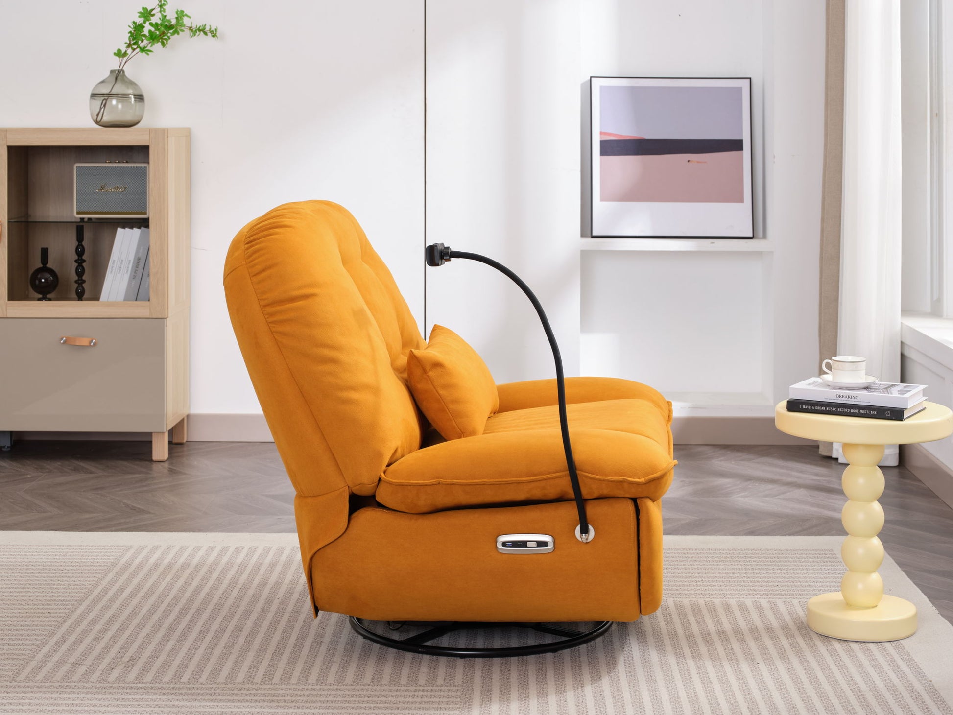 The Power Recliner Swivel Glider with USB charger and Bluetooth music player in mustard yellow, featuring a pillow and black reading light, sits next to a round side table with a cup and book in a modern living room with bookshelf and framed art.