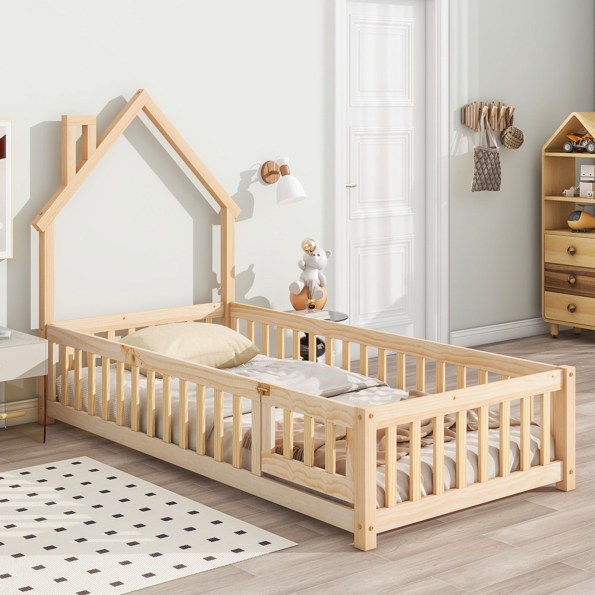 The House-Shaped Headboard Floor Bed With Fence features a wooden frame and safety rails, set in a bright, modern room with light walls, wood floors, cozy decor, a teddy bear, and shelves in the background.