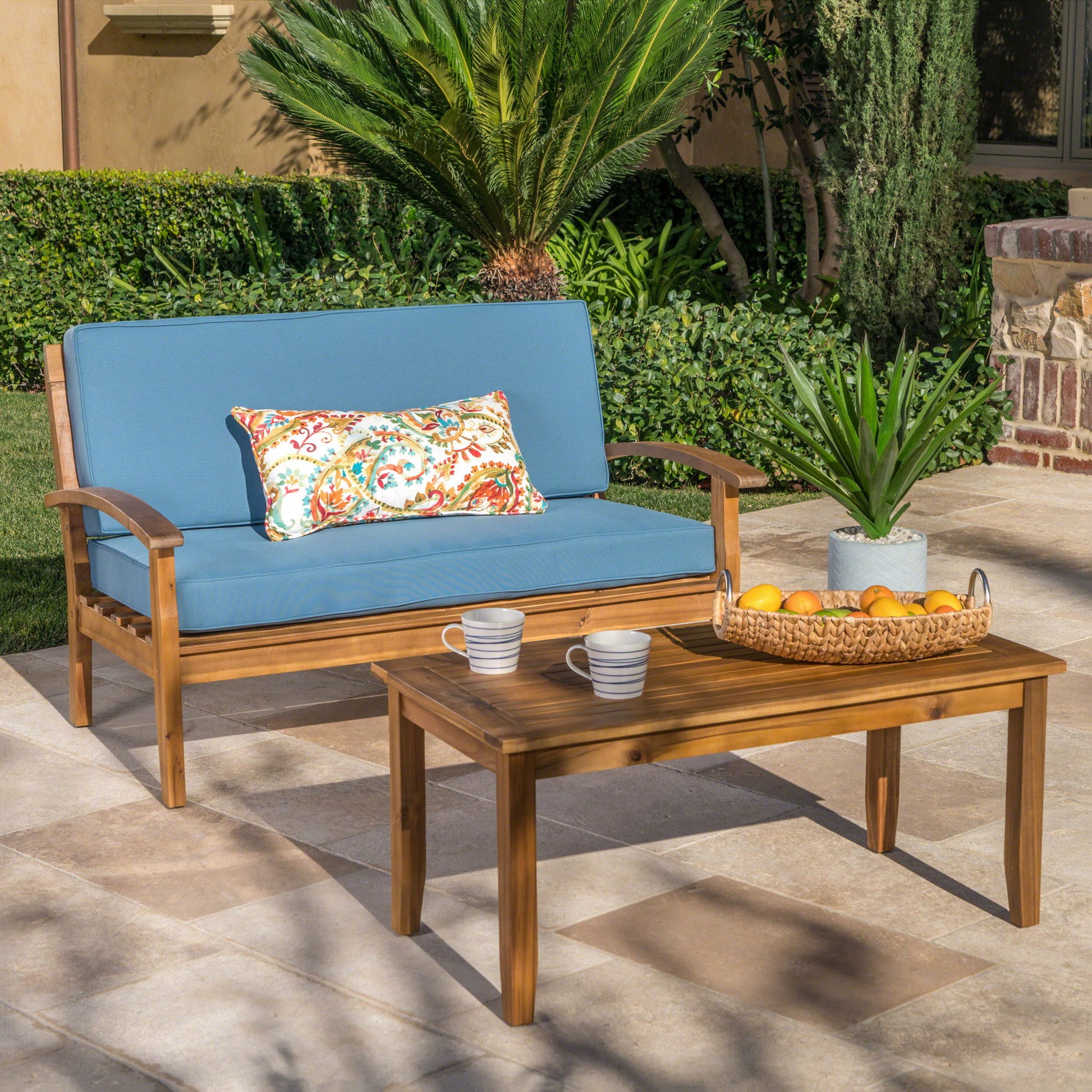 The Outdoor Chat Set With Coffee Table And Loveseat features blue cushions, a colorful pillow, and is styled with a basket of oranges, plant, and striped mugs on the table in a sunny garden patio setting.