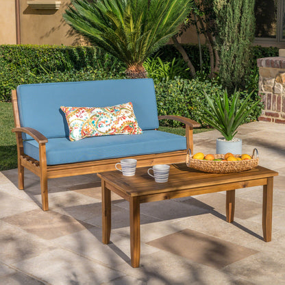 The Outdoor Chat Set With Coffee Table And Loveseat features blue cushions, a colorful pillow, and is styled with a basket of oranges, plant, and striped mugs on the table in a sunny garden patio setting.
