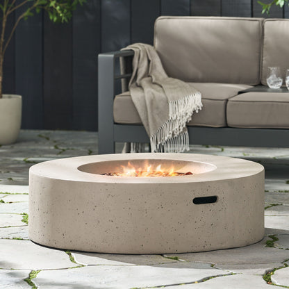 The Modern Lightweight Concrete Outdoor Circular Fire Pit 50,000 Btu sits on a stone patio beside a gray outdoor sofa with a fringed blanket over the armrest, its small central flame adding a contemporary touch.