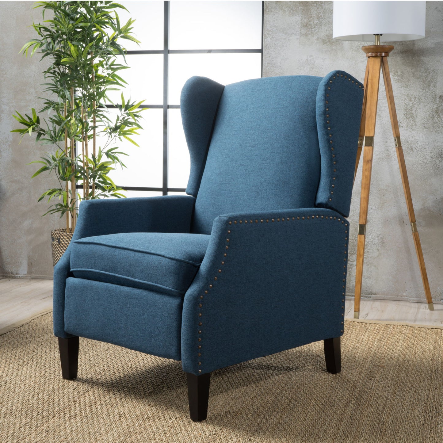 The Westeros Wing Back Fabric Upholstered Recliner with nailhead trim sits on a woven rug in a modern room, accented by a tall plant, floor lamp, and large windows in the background.