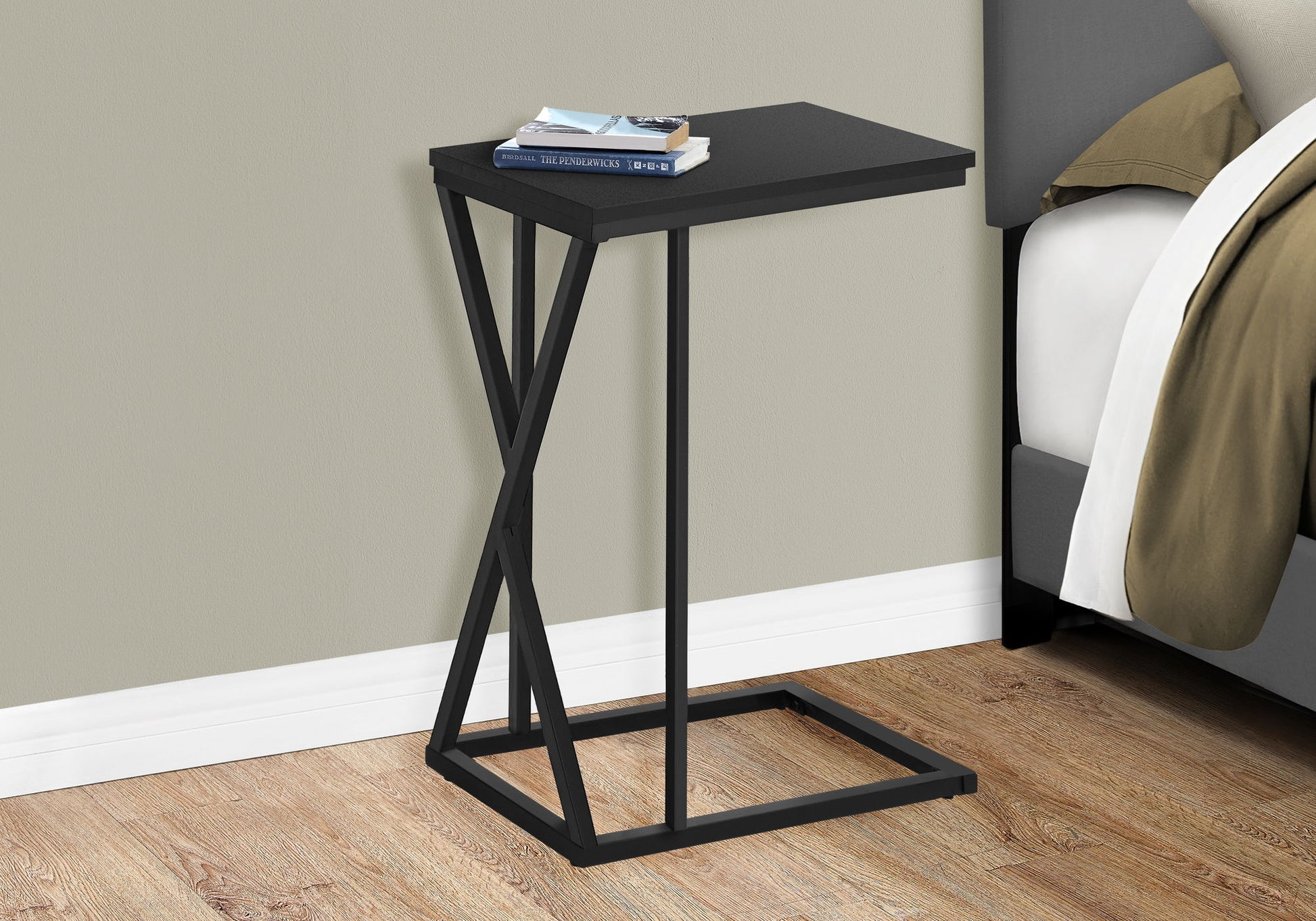 The Accent Table C - Shaped, Contemporary & Modern is a compact black metal side table with a rectangular top and geometric base, shown beside a bed holding two books and glasses, against a light wall and wood flooring.