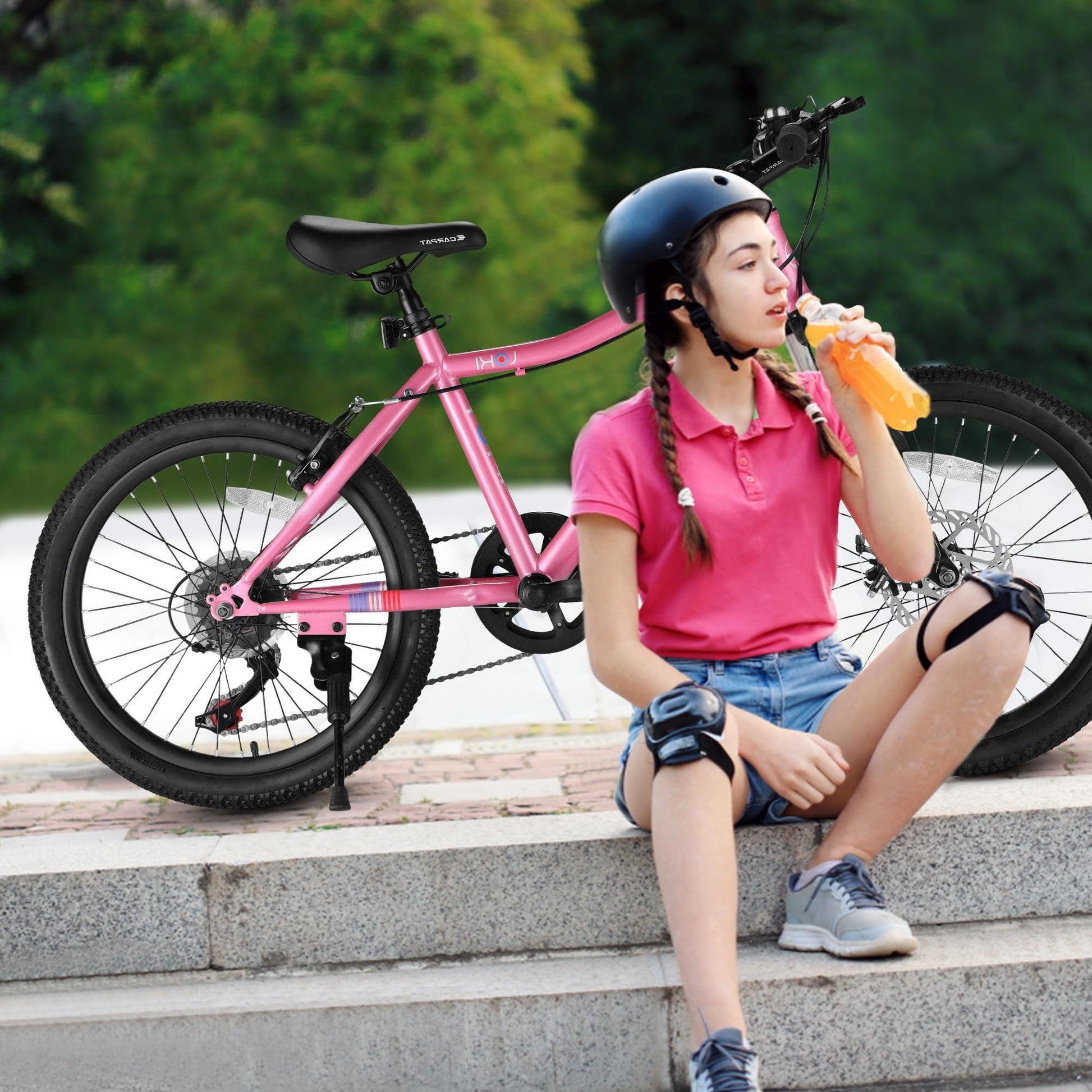 A young girl wearing a helmet, pink shirt, and knee pads sits on steps next to the S20101 20" Kids' Bike, drinking from an orange bottle in a green outdoor park setting.