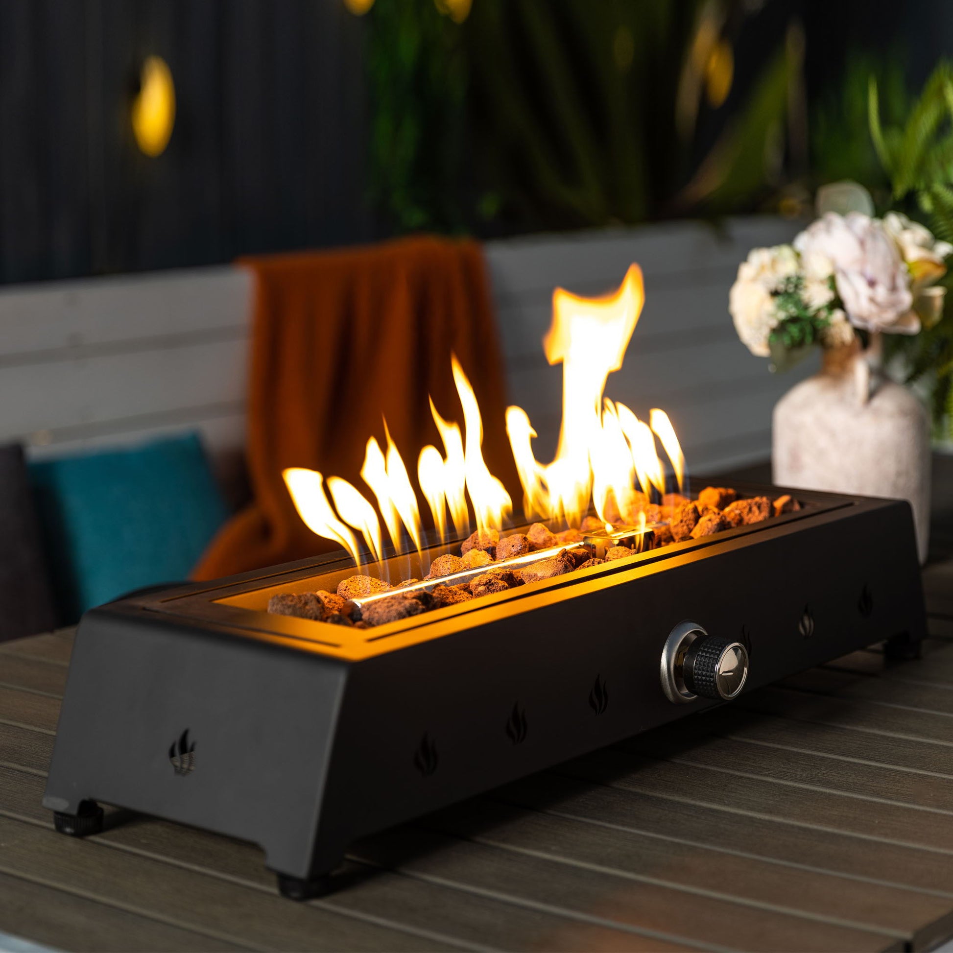 A 28" black tabletop propane gas fire pit with a glass wind guard and lava rocks sits on a wooden outdoor table. The portable fire pit features a front knob, with flowers in a vase and an orange blanket visible in the background.