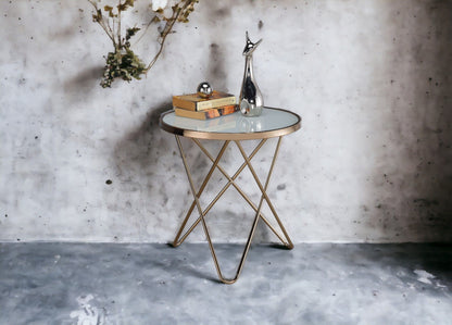 The Valora Glass Coffee Table with gold metal legs stands against a concrete wall. On it are two stacked books, a small silver sphere, and a tall abstract silver sculpture. Dried flowers hang on the wall above.