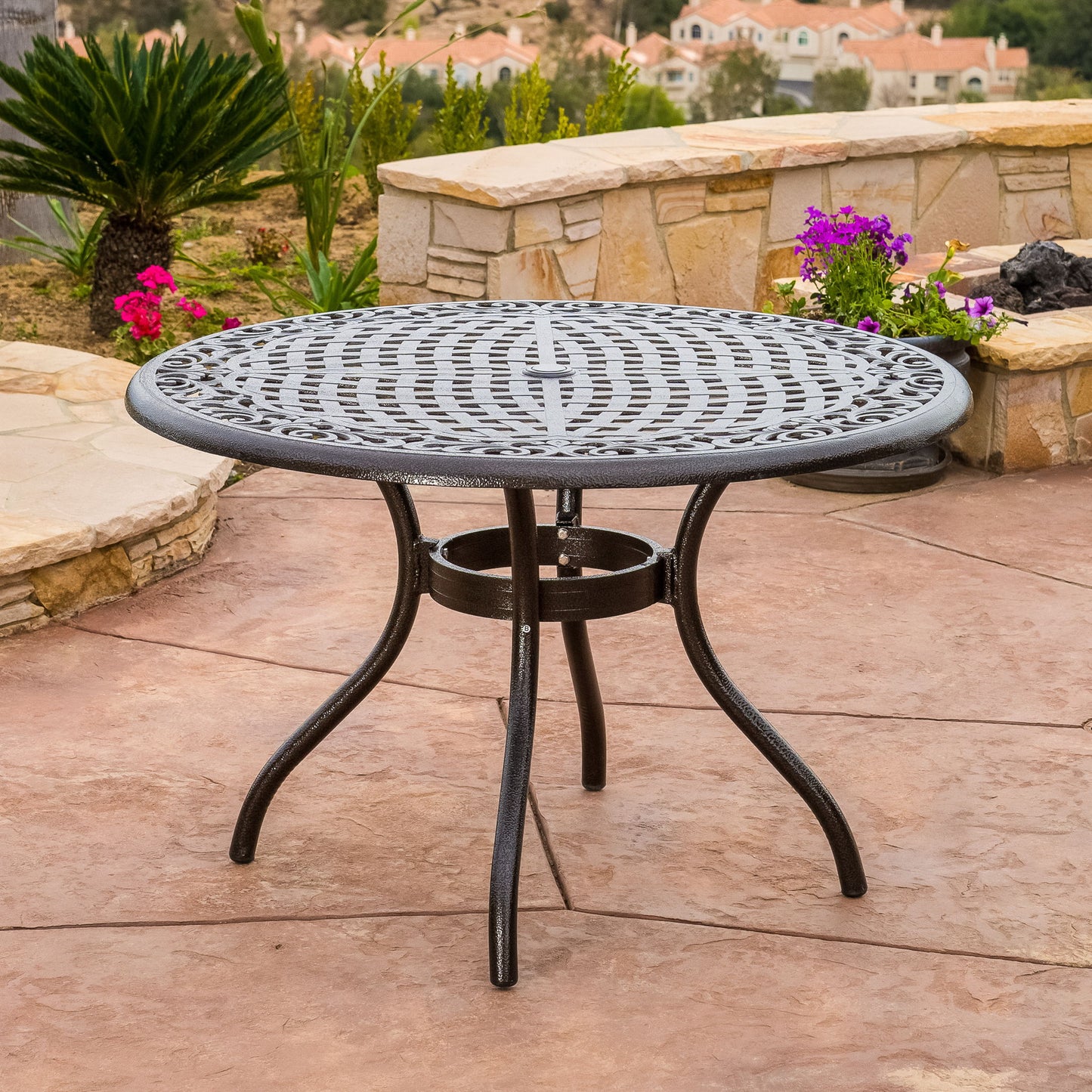 The Tranquilius Outdoor Round Dining Table, featuring a lattice tabletop, umbrella hole, and curved legs, stands on a stone patio surrounded by plants, flowers, and a stone wall with houses and greenery in the background.