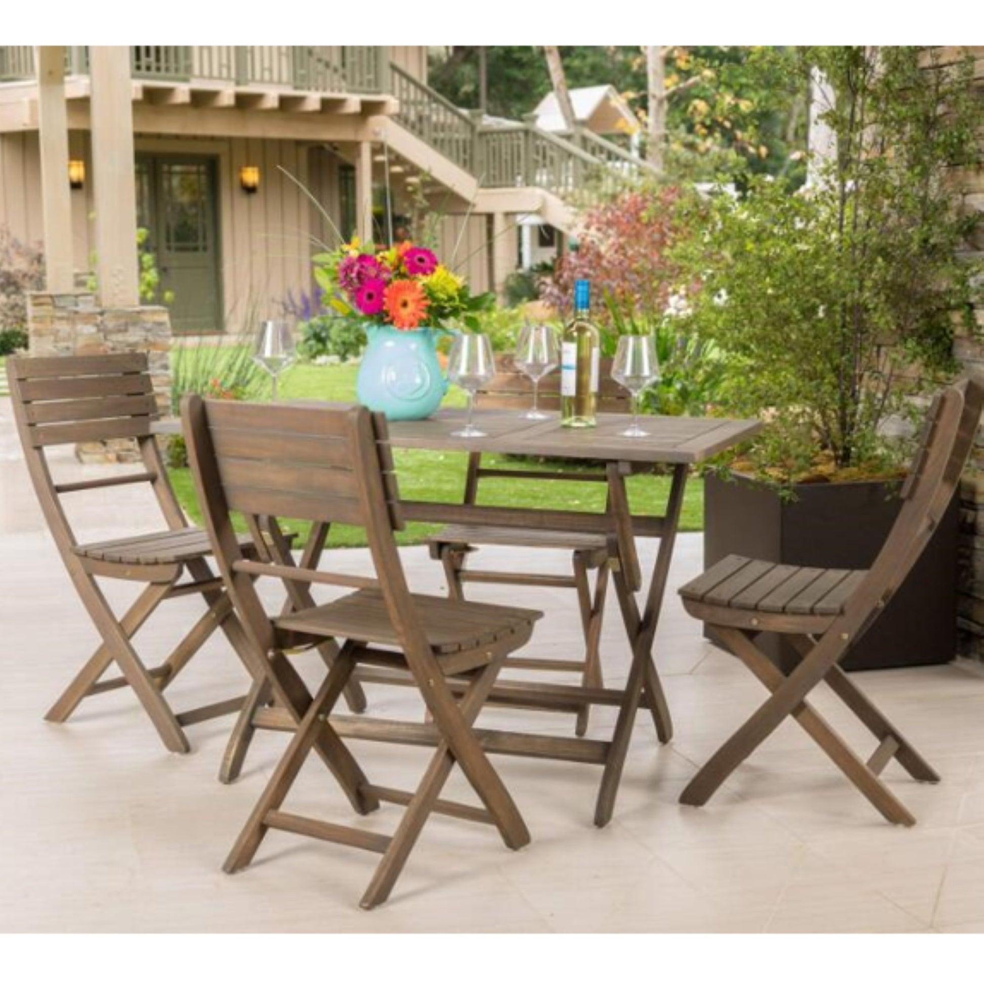 The Positano - Foldable Dining Set, featuring a square table and four chairs, is arranged outdoors with a vase of colorful flowers, wine glasses, and a wine bottle. Green plants and a house appear in the background.