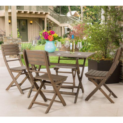 The Positano - Foldable Dining Set, featuring a square table and four chairs, is arranged outdoors with a vase of colorful flowers, wine glasses, and a wine bottle. Green plants and a house appear in the background.
