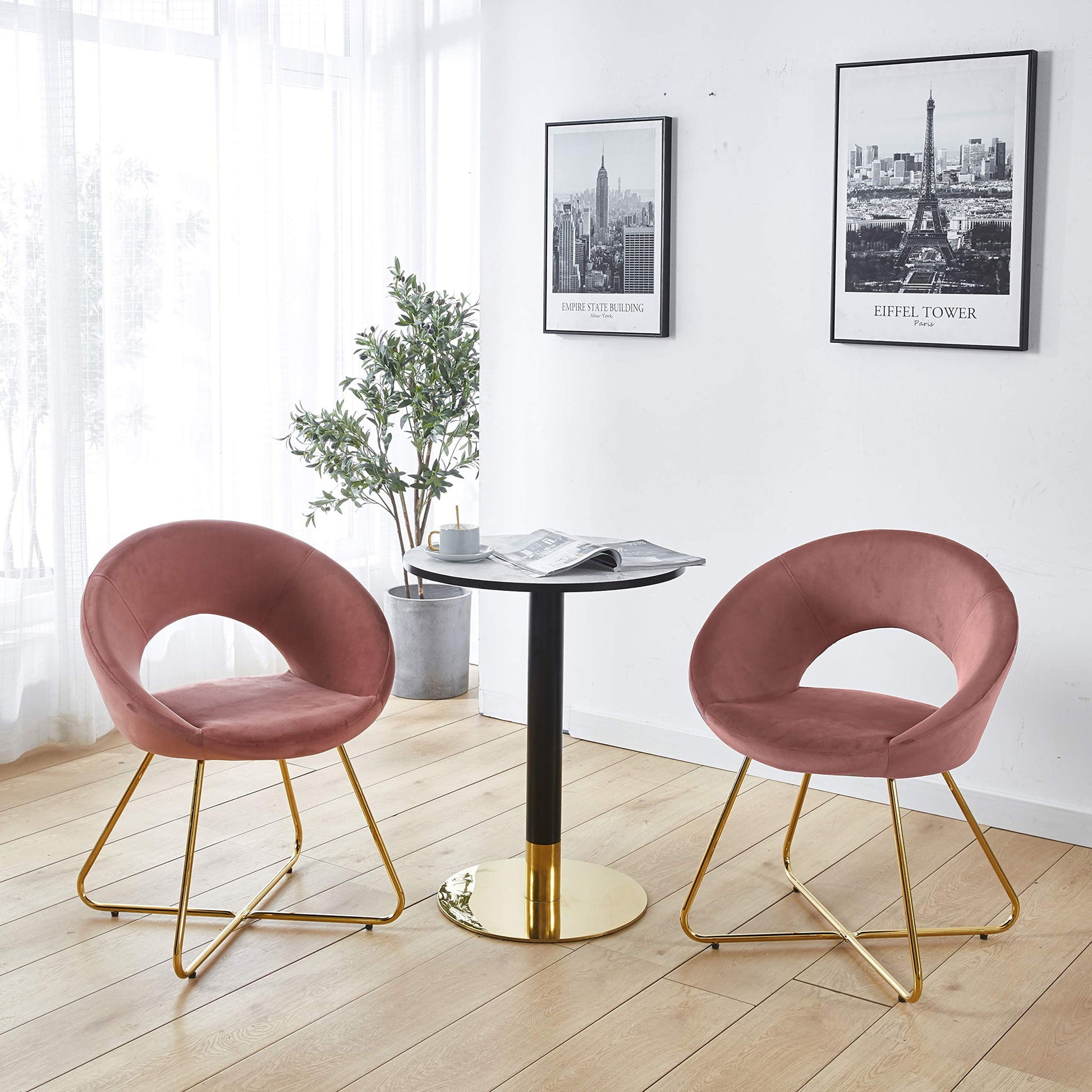 Two Modern Accent Velvet Lounge Chairs in pink with gold legs surround a small round table with a gold base. A newspaper rests on the table, while a potted plant and two framed cityscape photos decorate the white wall of this bright room.