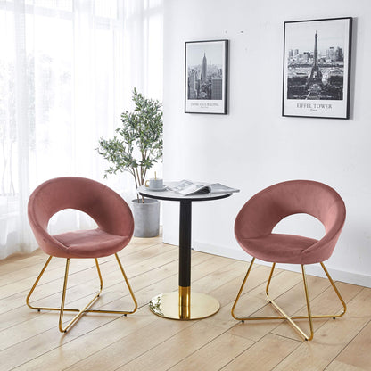 Two Modern Accent Velvet Lounge Chairs in pink with gold legs surround a small round table with a gold base. A newspaper rests on the table, while a potted plant and two framed cityscape photos decorate the white wall of this bright room.