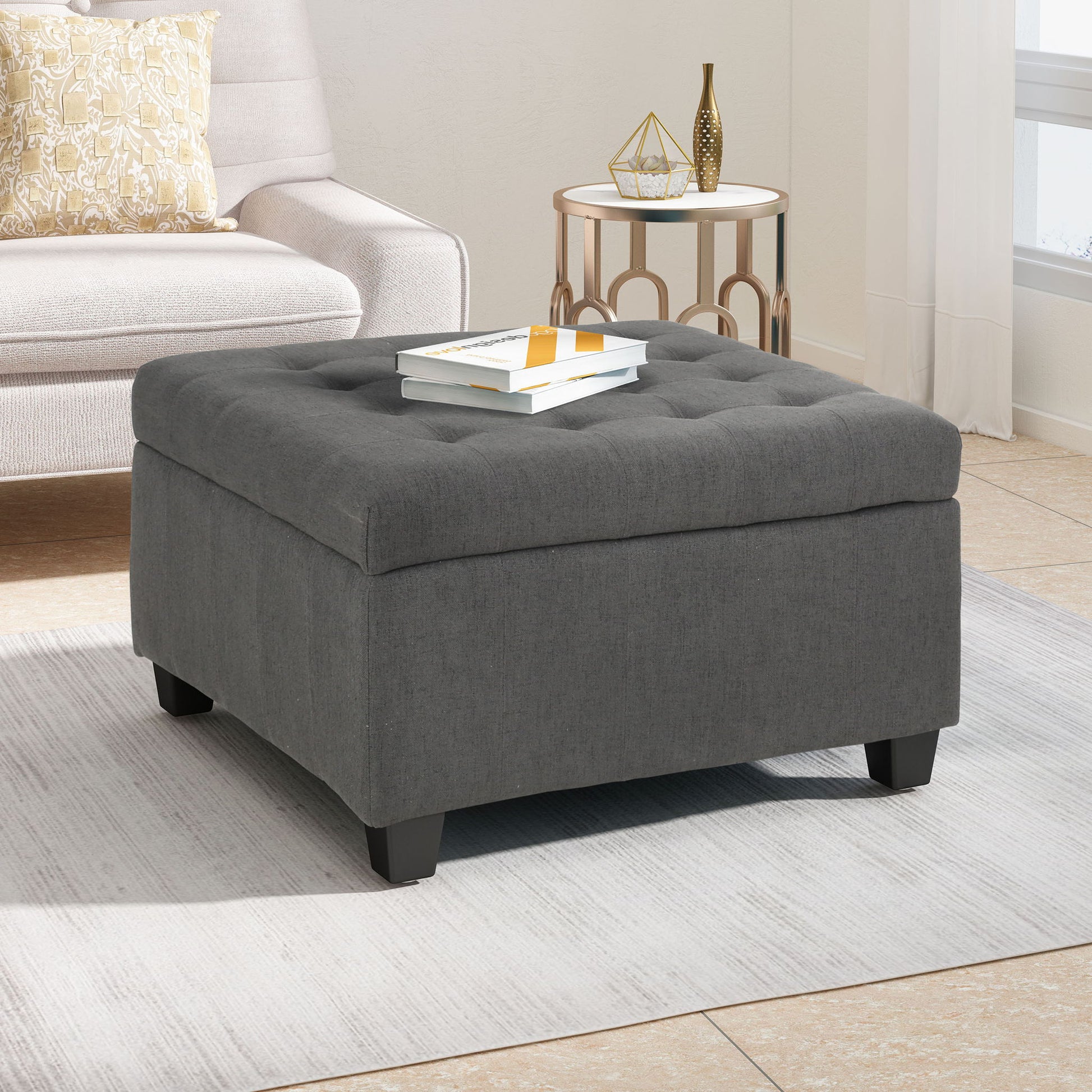 The Isabella Contemporary Ottoman With Storage in gray sits on a light area rug in a bright living room, topped with two books. A beige sofa with a yellow patterned pillow and a small round side table appear in the background.