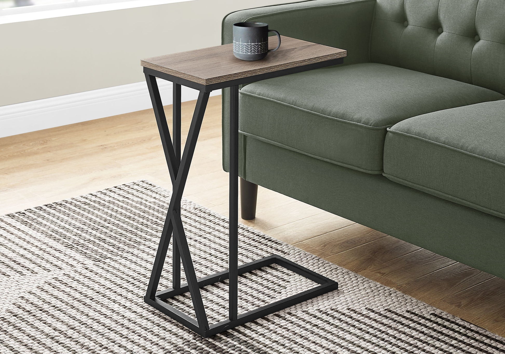 The Accent Table C-Shaped, Contemporary & Modern with a wood top and black metal frame sits beside a green sofa on a striped rug, holding a black mug.