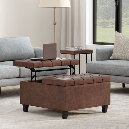 The Harrison Large Coffee Table Storage Ottoman with a lift-top tray sits between two gray sofas in the living room. Nearby, a laptop, lamp, and small table with books are visible by a large window overlooking trees.