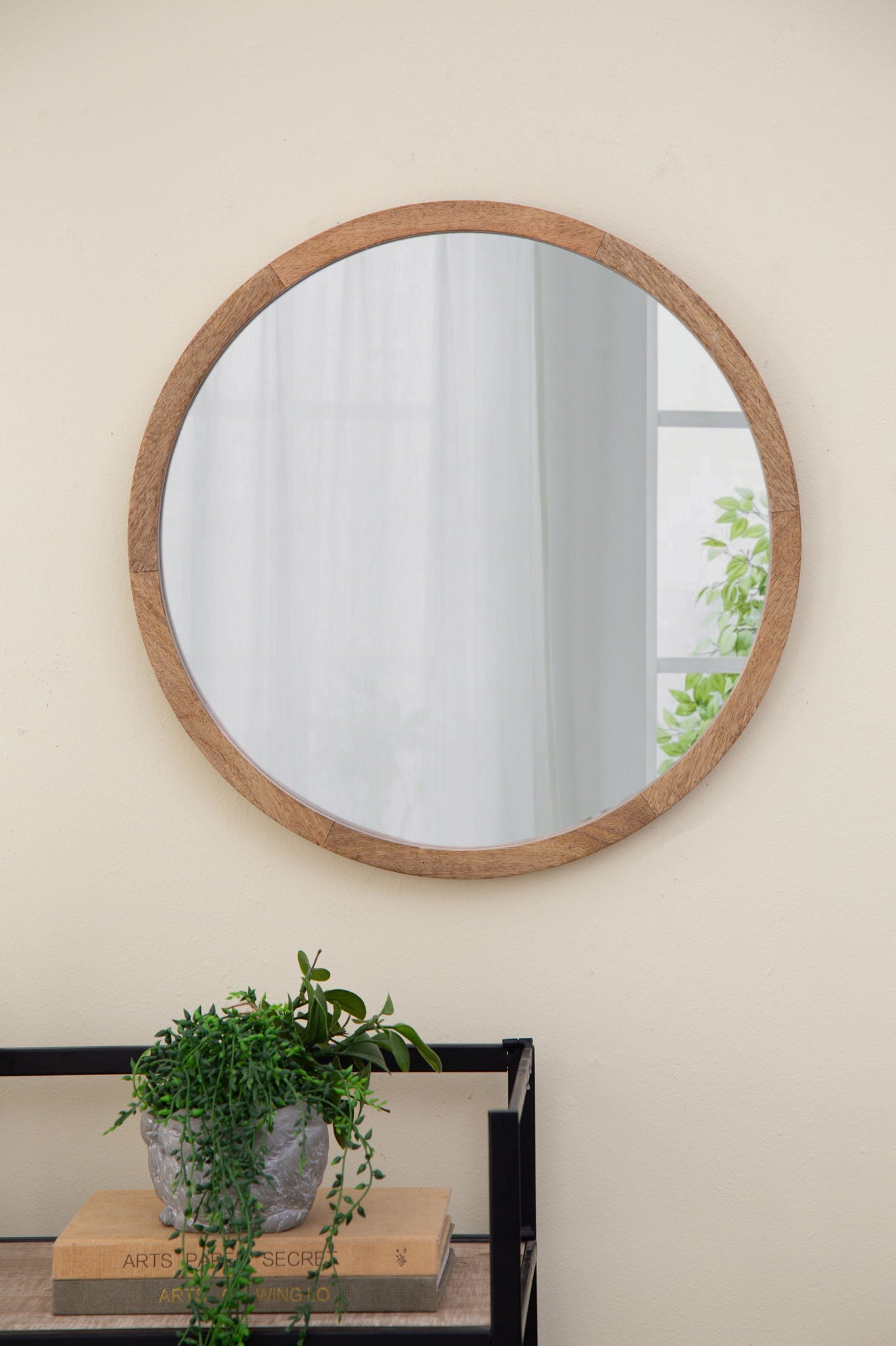 The Circle Wall Mirror With Wooden Frame hangs on a light-colored wall above a black metal shelf holding a potted plant and books, reflecting a window and greenery from outside. Ideal wall mirror for living room decor.