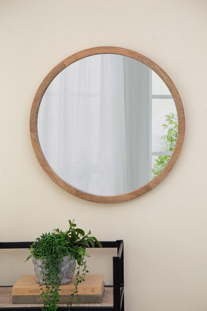 The Circle Wall Mirror With Wooden Frame hangs on a light-colored wall above a black metal shelf holding a potted plant and books, reflecting a window and greenery from outside. Ideal wall mirror for living room decor.