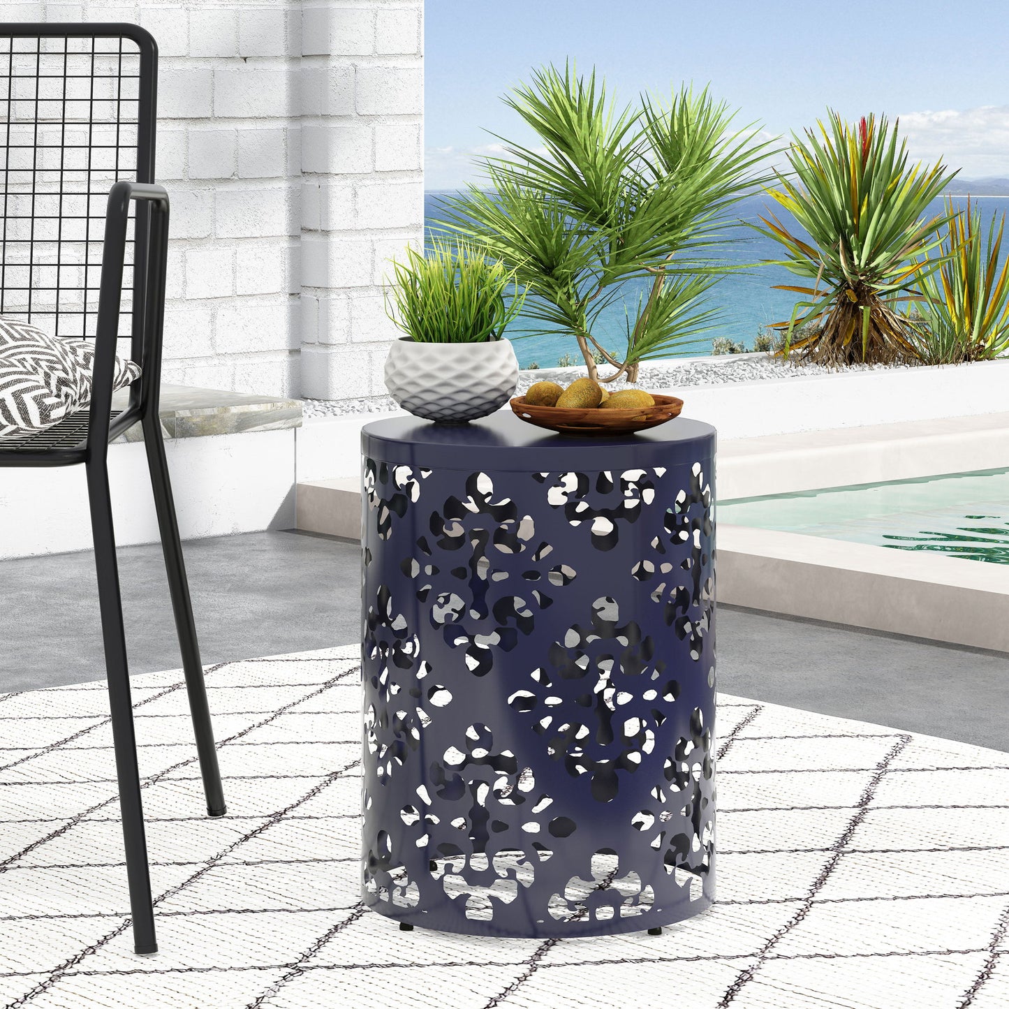 An Outdoor Metal Modern Design End Table in navy blue with cutout patterns holds two potted plants and a bowl of figs beside a chair, placed on a patterned rug by the pool with ocean views and palm plants in the background.