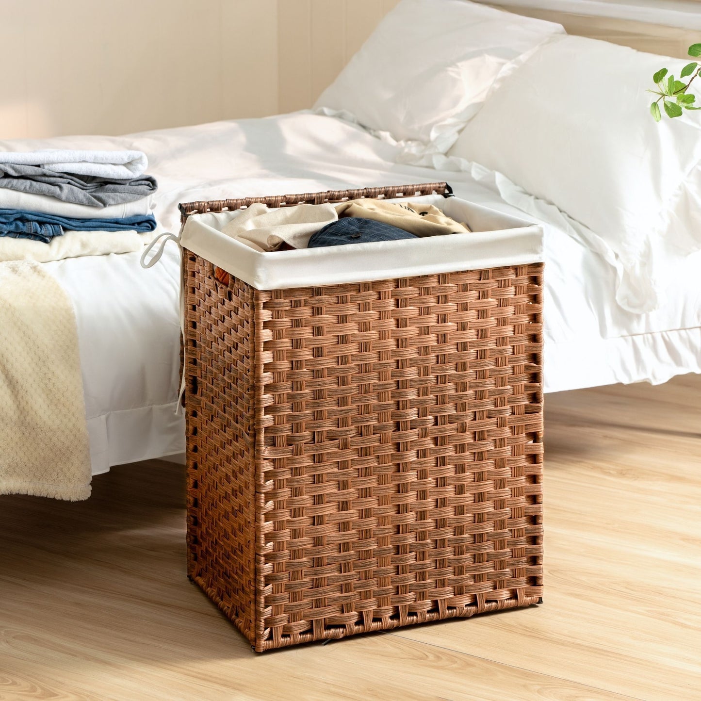 A Laundry Hamper With Lid PE Rattan Powder Coating Frame Clothes Hampers With 2 Removable Bags sits by a bed with white bedding; clothes peek from the hamper, and folded laundry rests on the bed.
