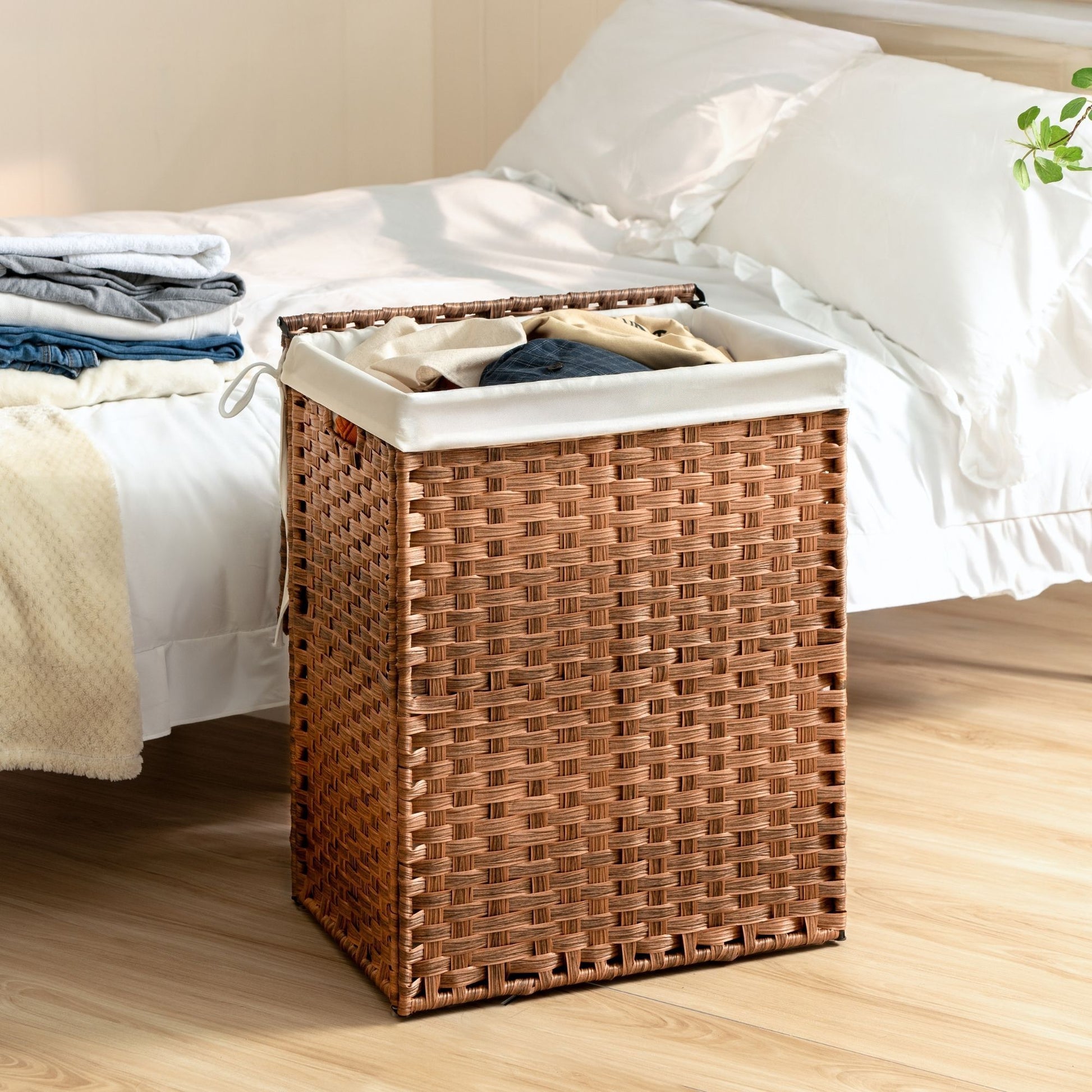 A Laundry Hamper With Lid PE Rattan Powder Coating Frame Clothes Hampers With 2 Removable Bags sits by a bed with white bedding; clothes peek from the hamper, and folded laundry rests on the bed.