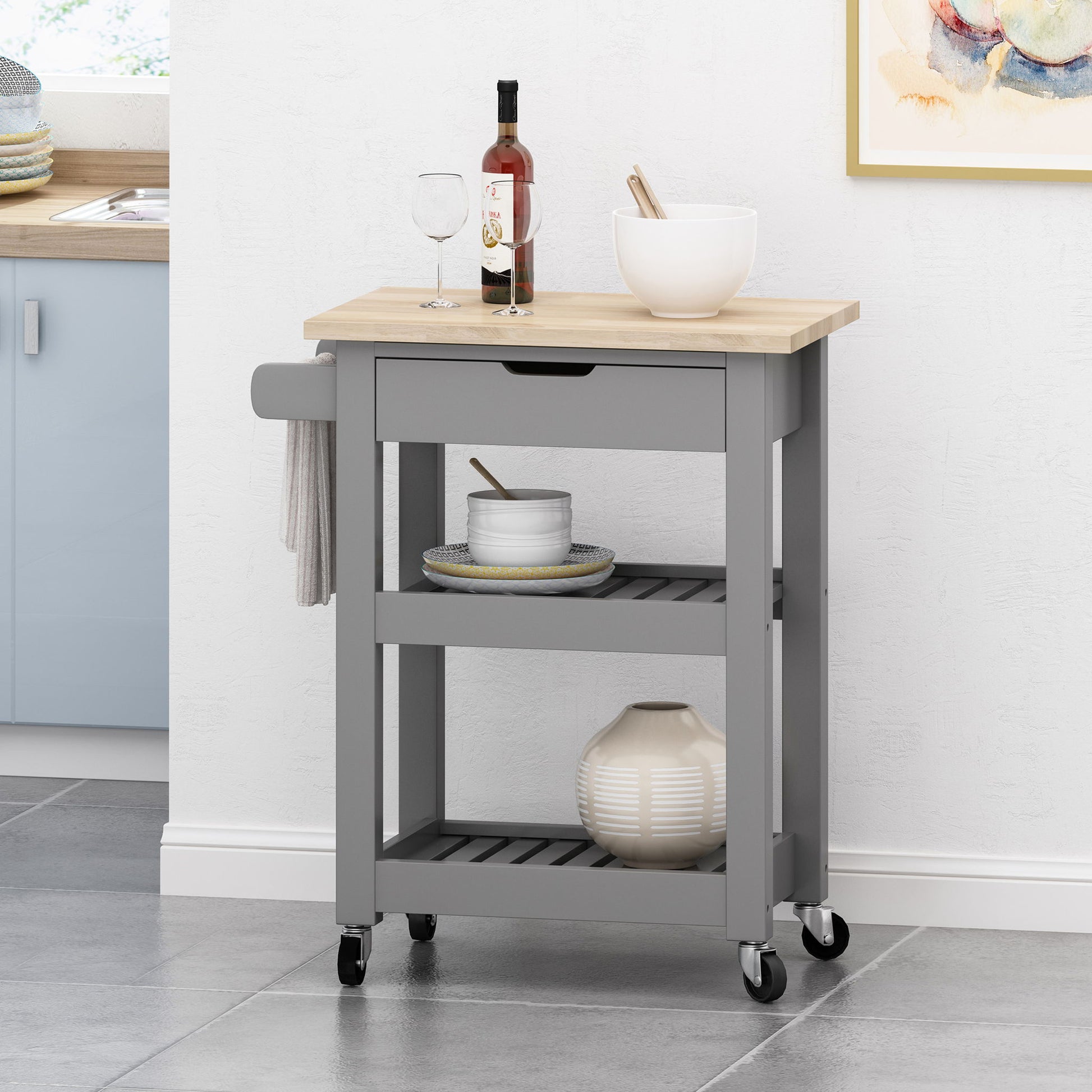 The Tattnall Kitchen Cart With Wheels features a compact gray frame, light wood top, two shelves, a side towel bar, storage for kitchen items, and four black wheels—perfect beside the counter on a tiled floor.