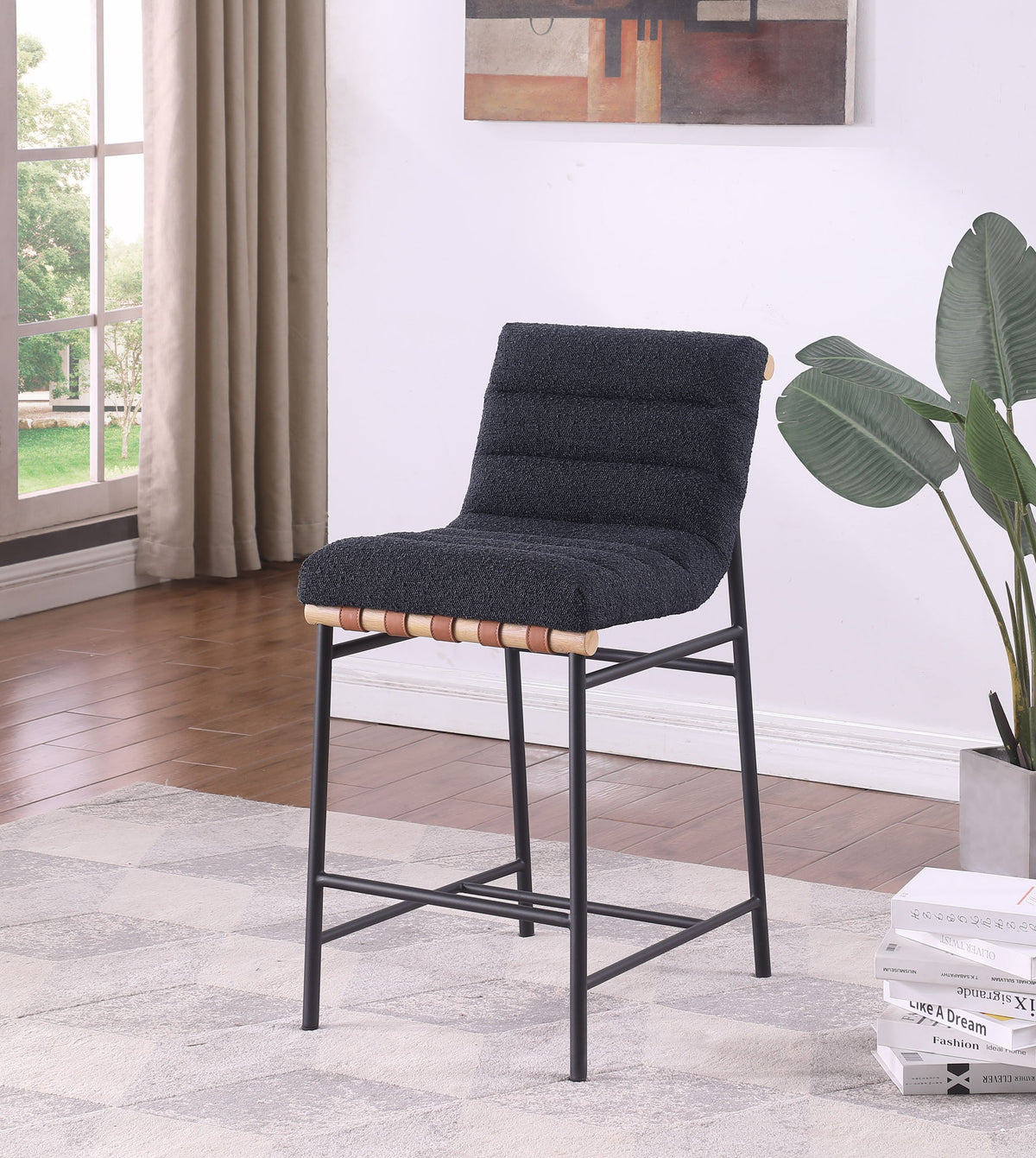 The Lahni - 17" Boucle Fabric Counter Height Chair with a modern black cushioned seat and metal frame stands on a light patterned rug in a bright room featuring wooden floors, a large window with beige curtains, a potted plant, and abstract wall art.