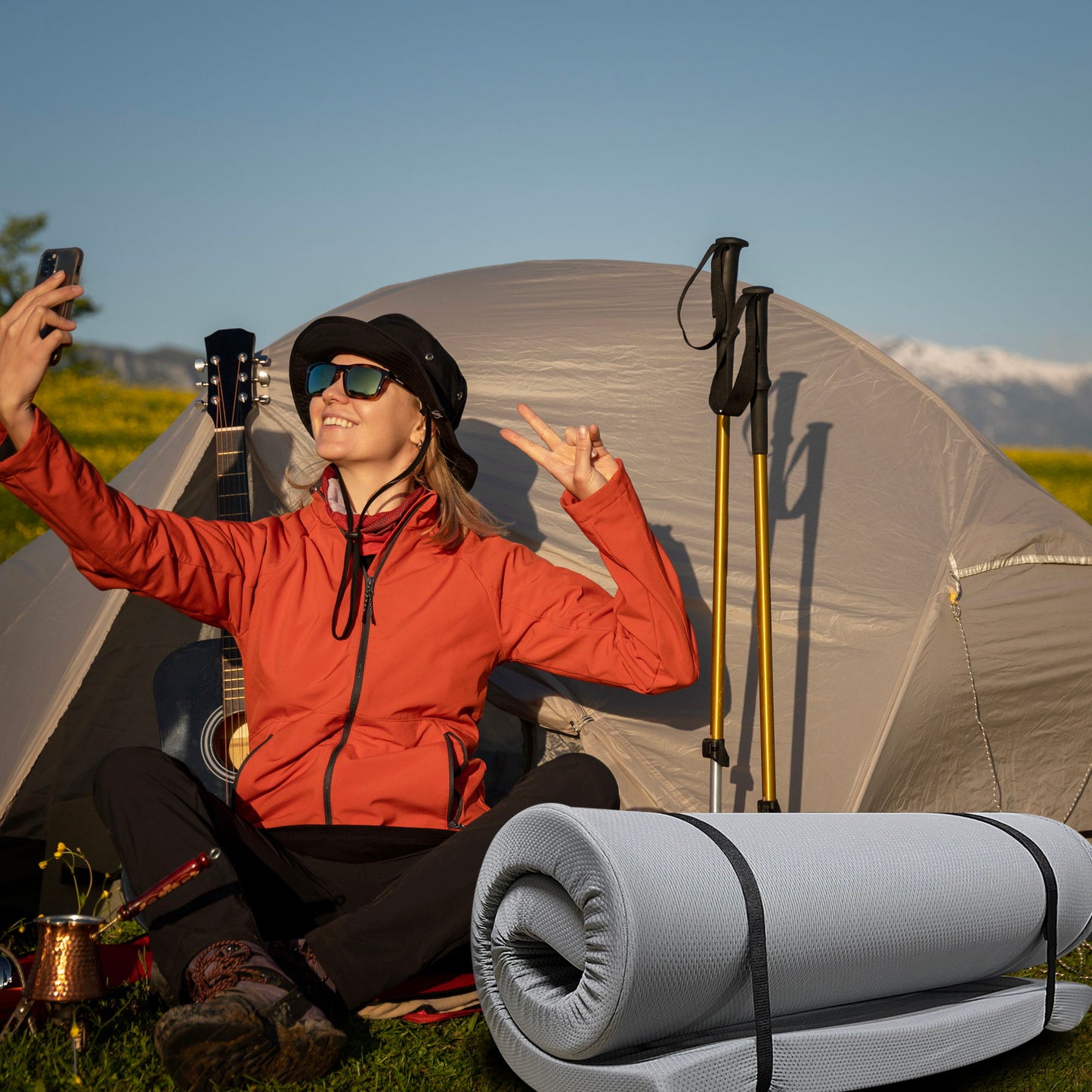 A woman in sunglasses and an orange jacket sits on grass by a tent, taking a selfie with a peace sign. Nearby is the gray Memory Foam Camping Mattress Foldable, portable with water-resistant cover; mountains rise in the background.