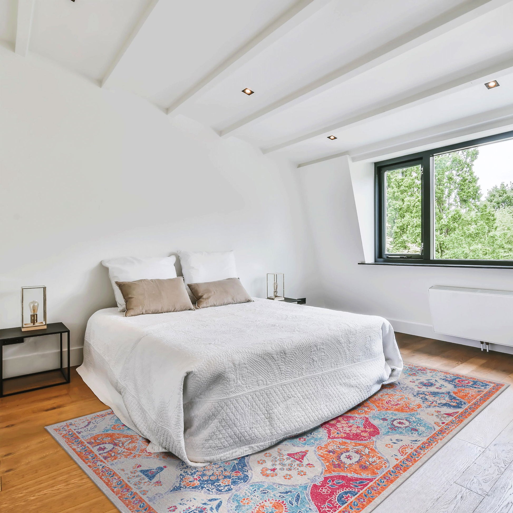 A bright, modern bedroom features a white bed with two beige pillows, minimalist black nightstands, and the Maval - Area Rug adding color to wooden flooring. Large windows fill the room with natural light and reveal green trees outside.