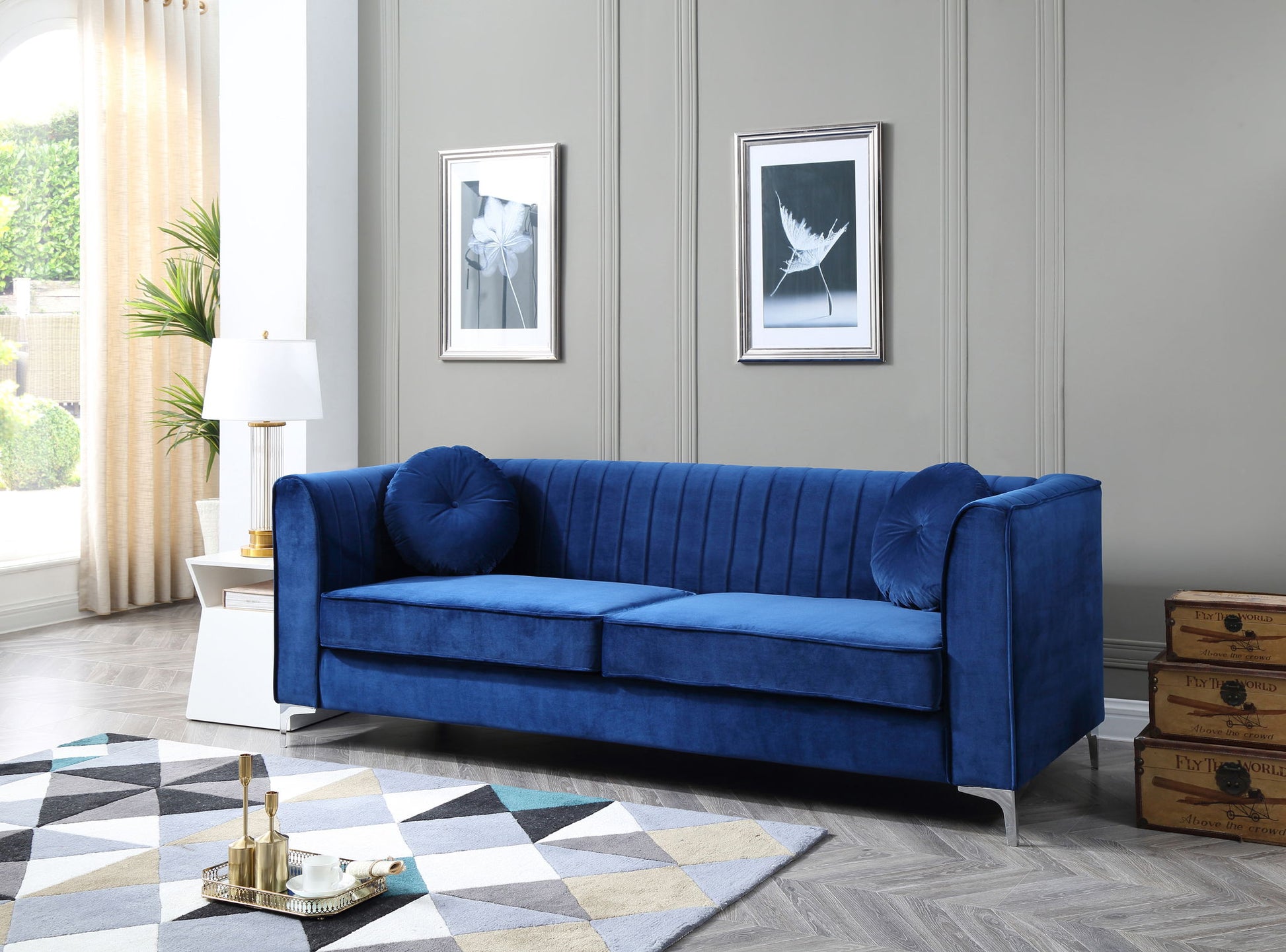 A modern living room features the Delray Micro Suede Sofa in blue with two round cushions, a white side table topped by a gold lamp, framed art on gray walls, a geometric rug, and stacked vintage suitcases in the corner.