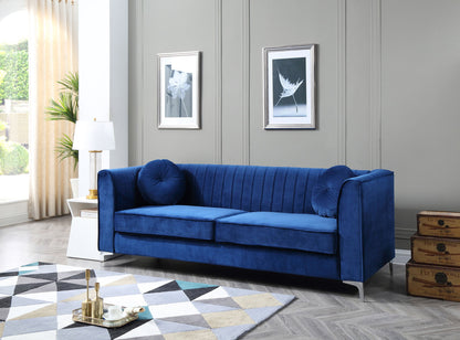 A modern living room features the Delray Micro Suede Sofa in blue with two round cushions, a white side table topped by a gold lamp, framed art on gray walls, a geometric rug, and stacked vintage suitcases in the corner.