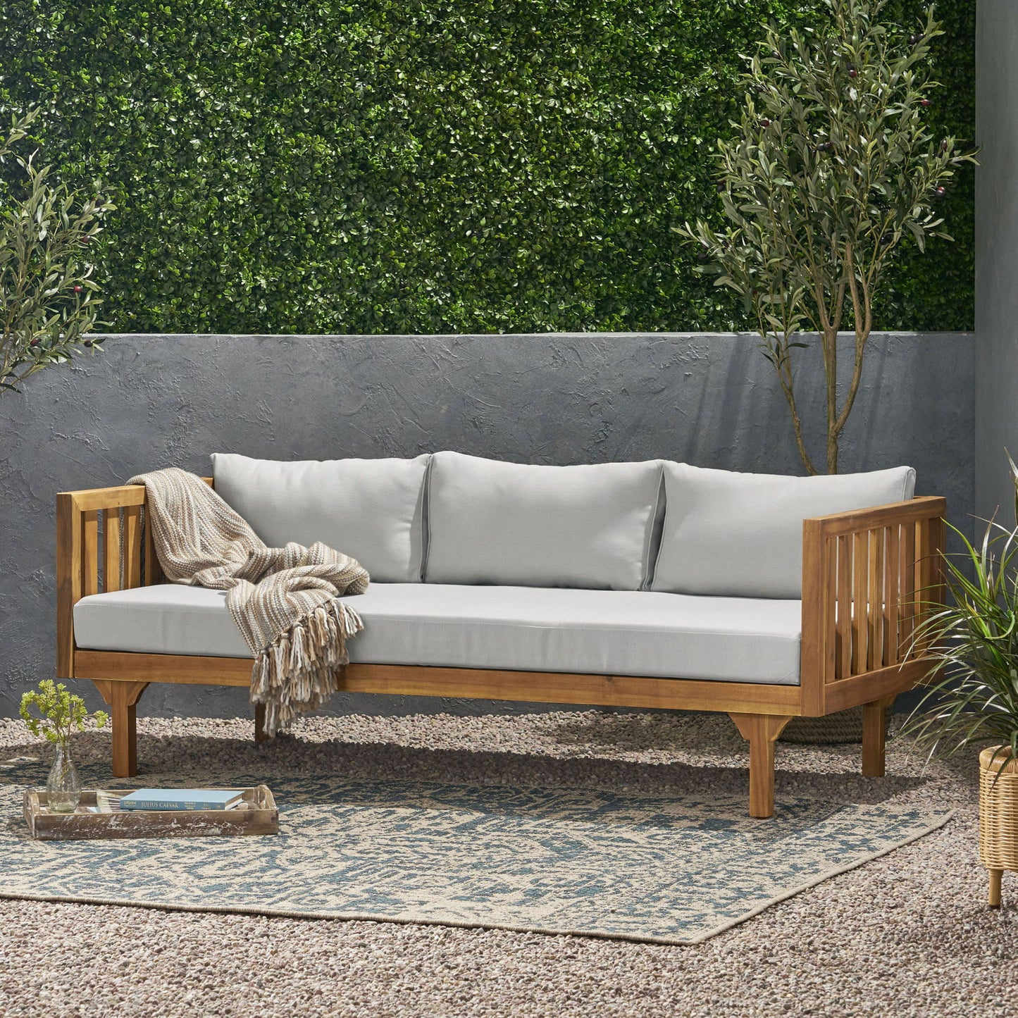 The Claremont 3 Seater Daybed, made from teak and acacia wood with water-resistant cushions, sits on a patterned rug in a gravel garden, accented by a beige throw blanket and surrounded by lush plants and a tall hedge.