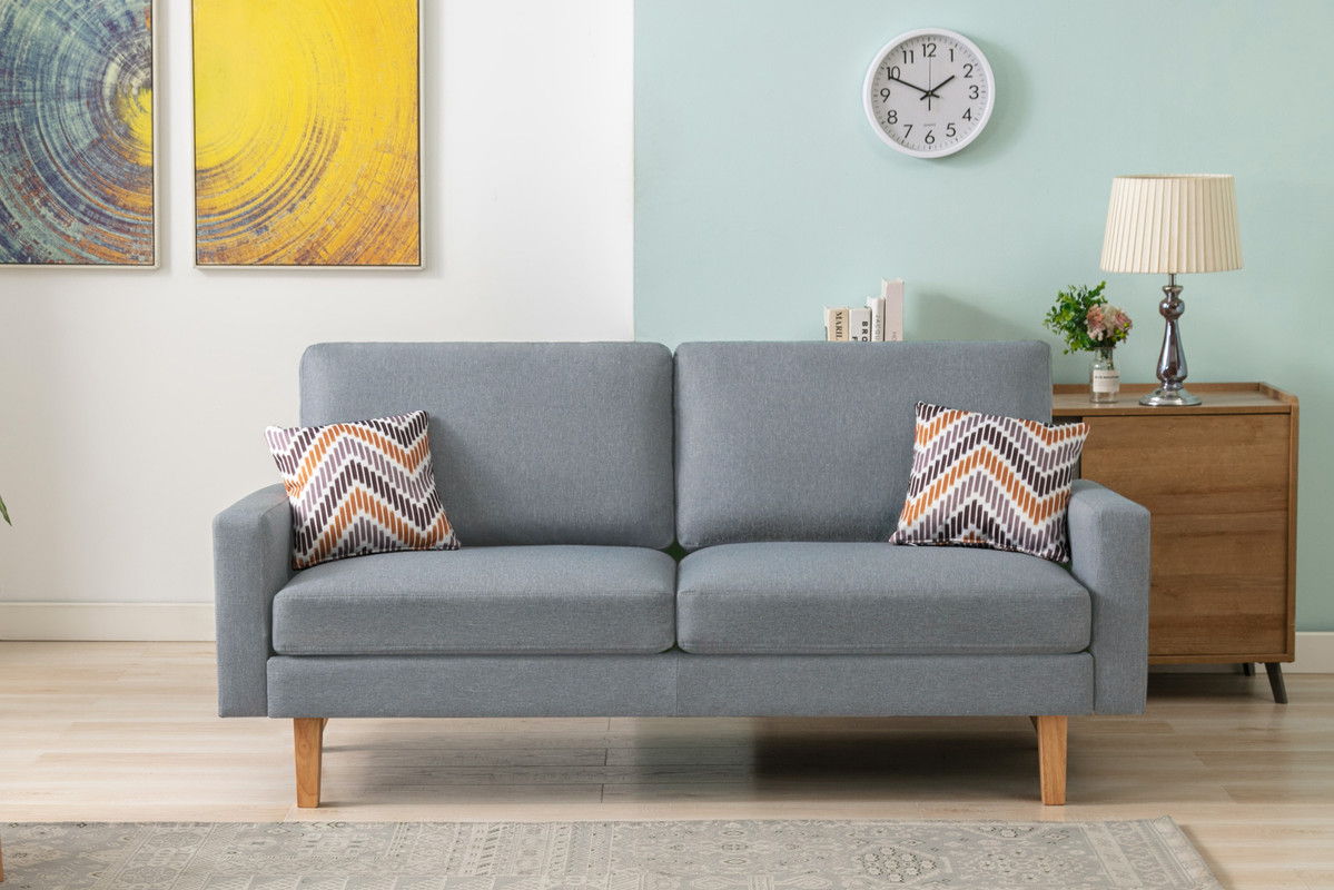 A modern living room featuring the Bahamas Sofa Set with 2 throw pillows, a wooden side table with a lamp and plant, wall art, a clock, and a decorative rug on a light wood floor.