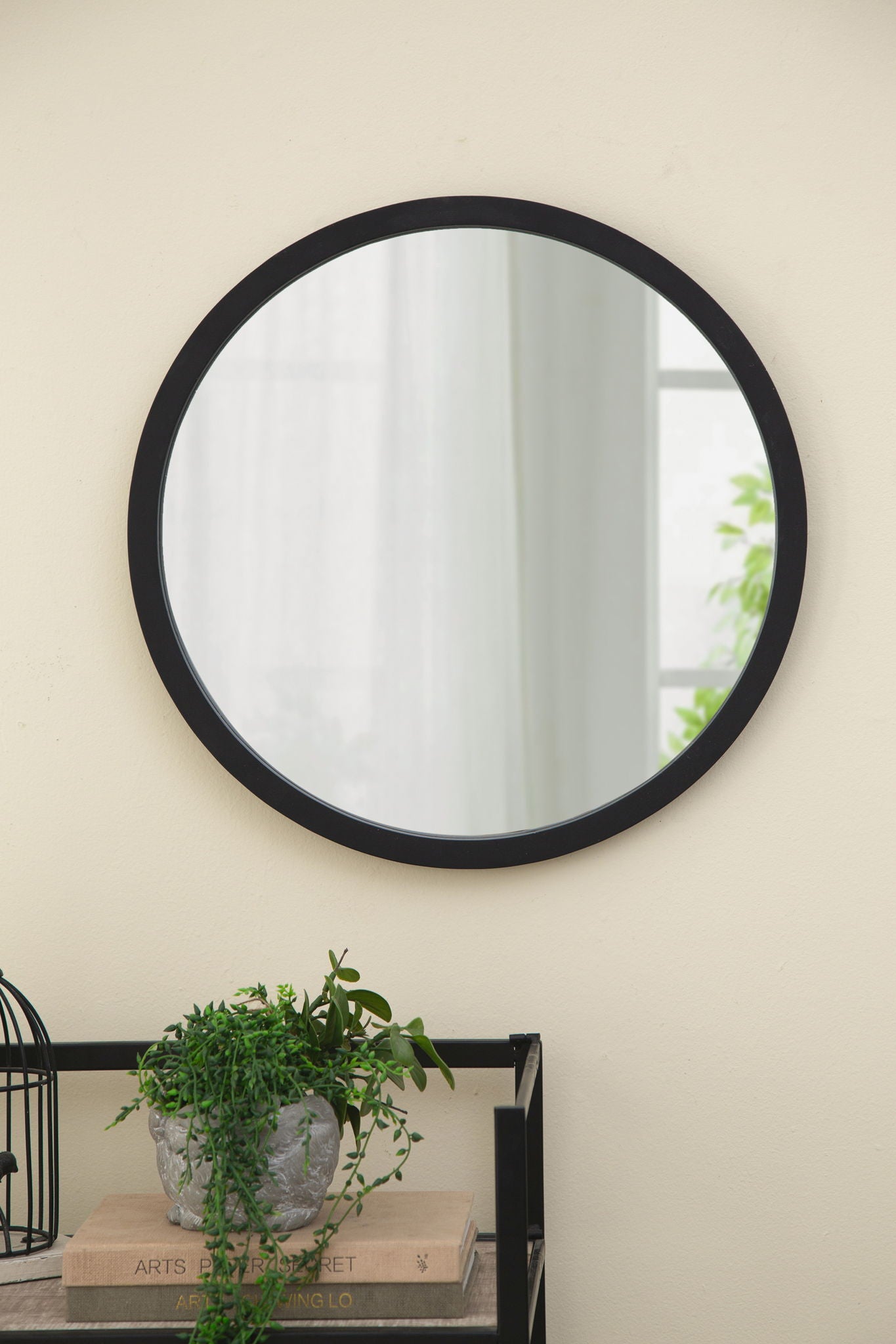 A Circle Wall Mirror With Wooden Frame hangs on a beige wall above a small table with a potted green plant, two stacked books, and part of a black wire decorative item.