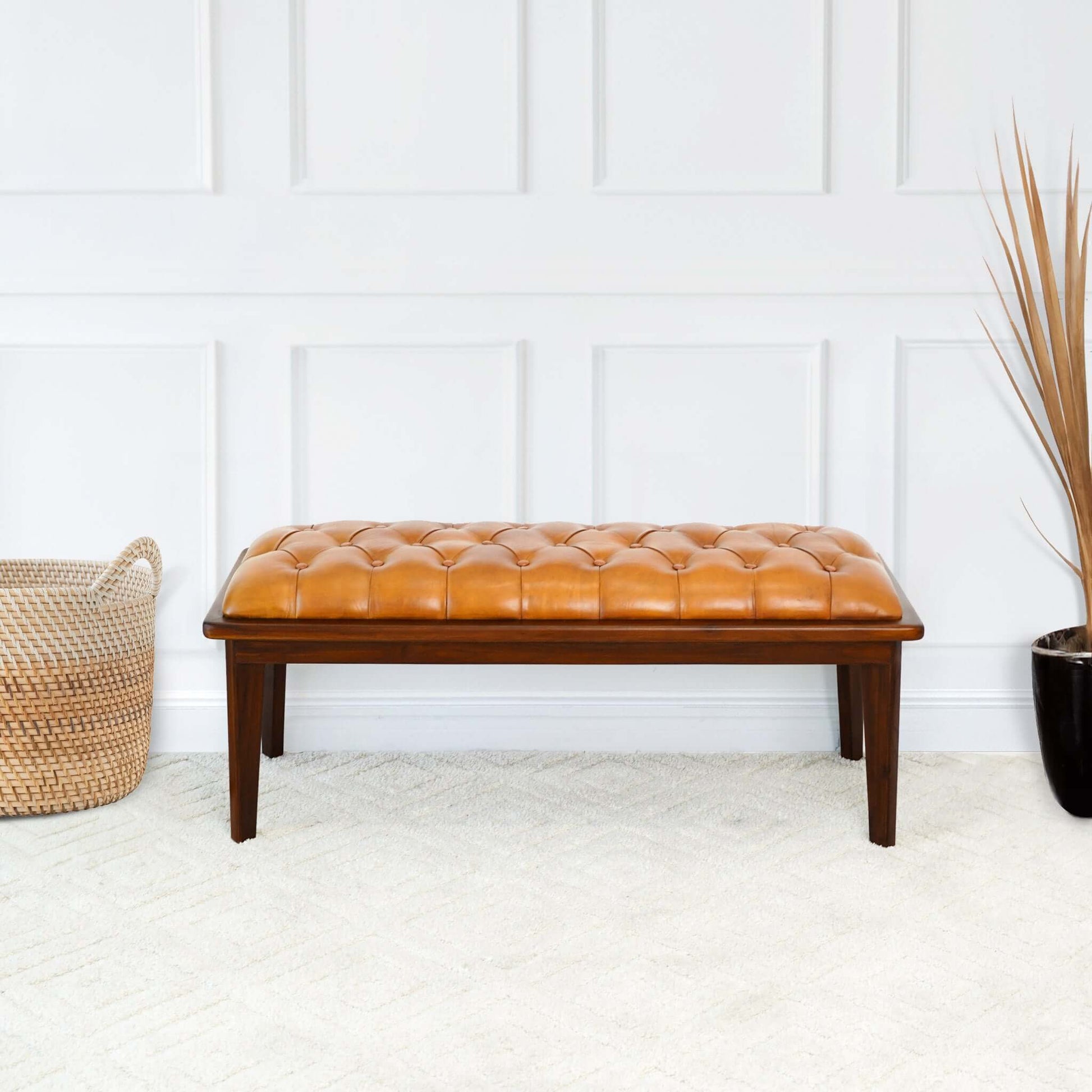 The Arden - Bench With Buttons in tan sits on a light carpet before a white paneled wall, flanked by a woven basket on the left and a black planter holding dried grass on the right.