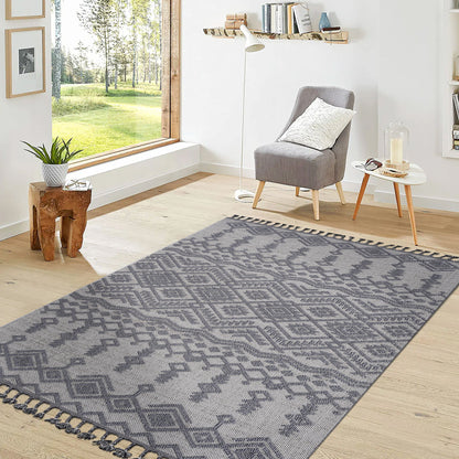 A modern living room with a large window, light wood floors, a Guros Traditional Indoor/Outdoor Area Rug in gray, a gray armchair with a white pillow, small round table, wooden stool, and wall shelves with books and decor.