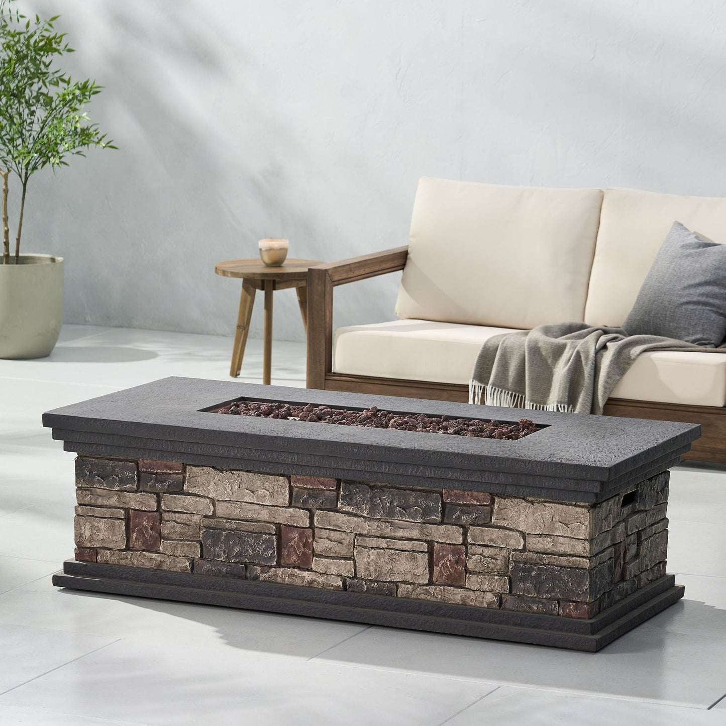 A Fire Pit With Lightweight Concrete And Steel Frame, featuring a recessed area with lava rocks, sits in front of a cushioned wooden sofa draped with a gray throw and accompanied by a small side table holding a candle.
