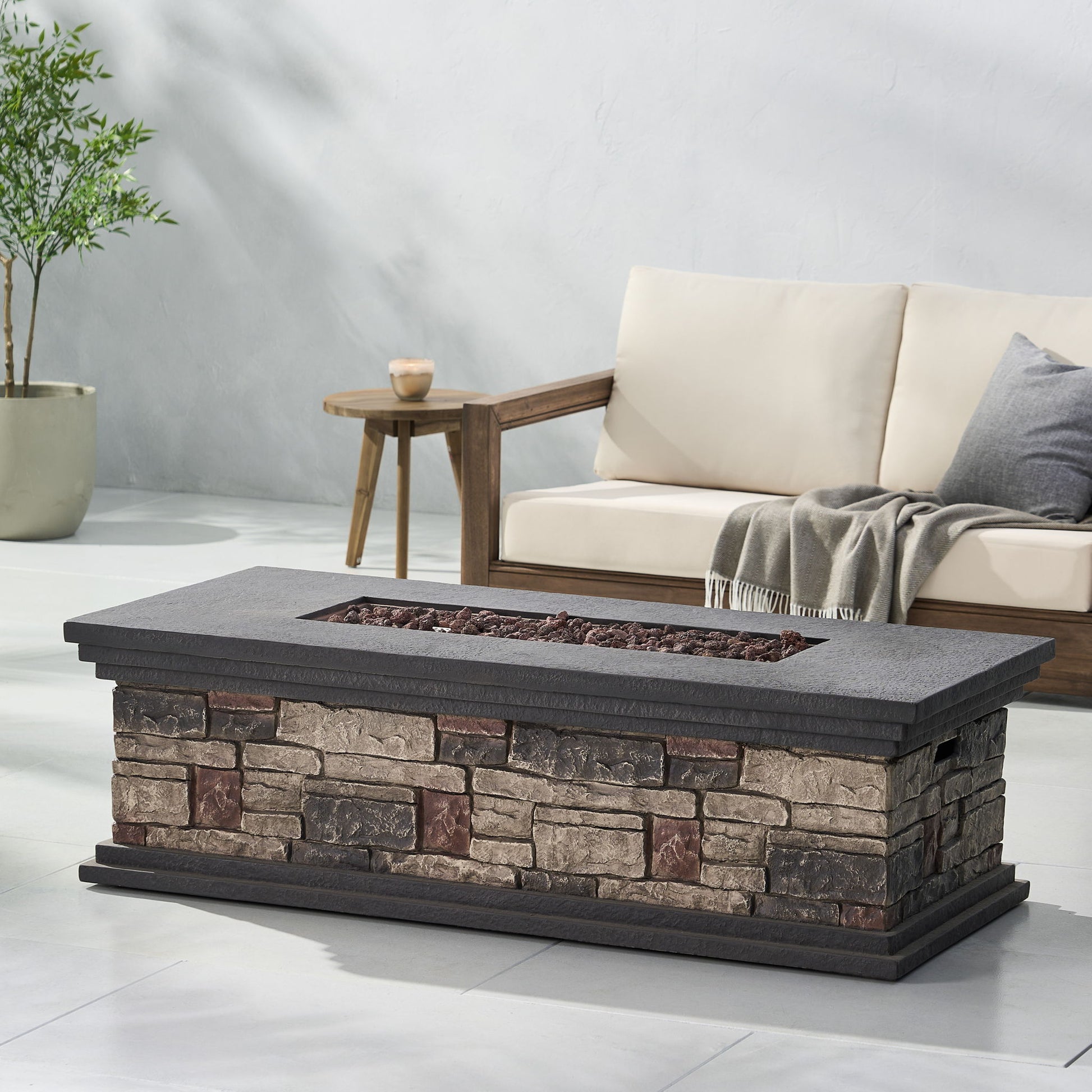 A Fire Pit With Lightweight Concrete And Steel Frame, featuring a recessed area with lava rocks, sits in front of a cushioned wooden sofa draped with a gray throw and accompanied by a small side table holding a candle.