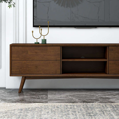 The Caroline - Modern TV Stand Up To 72" features angled legs, two drawers, and open shelves. It sits on a gray marble floor with a gold candelabra and small plant on top, while a flat-screen TV is mounted above.