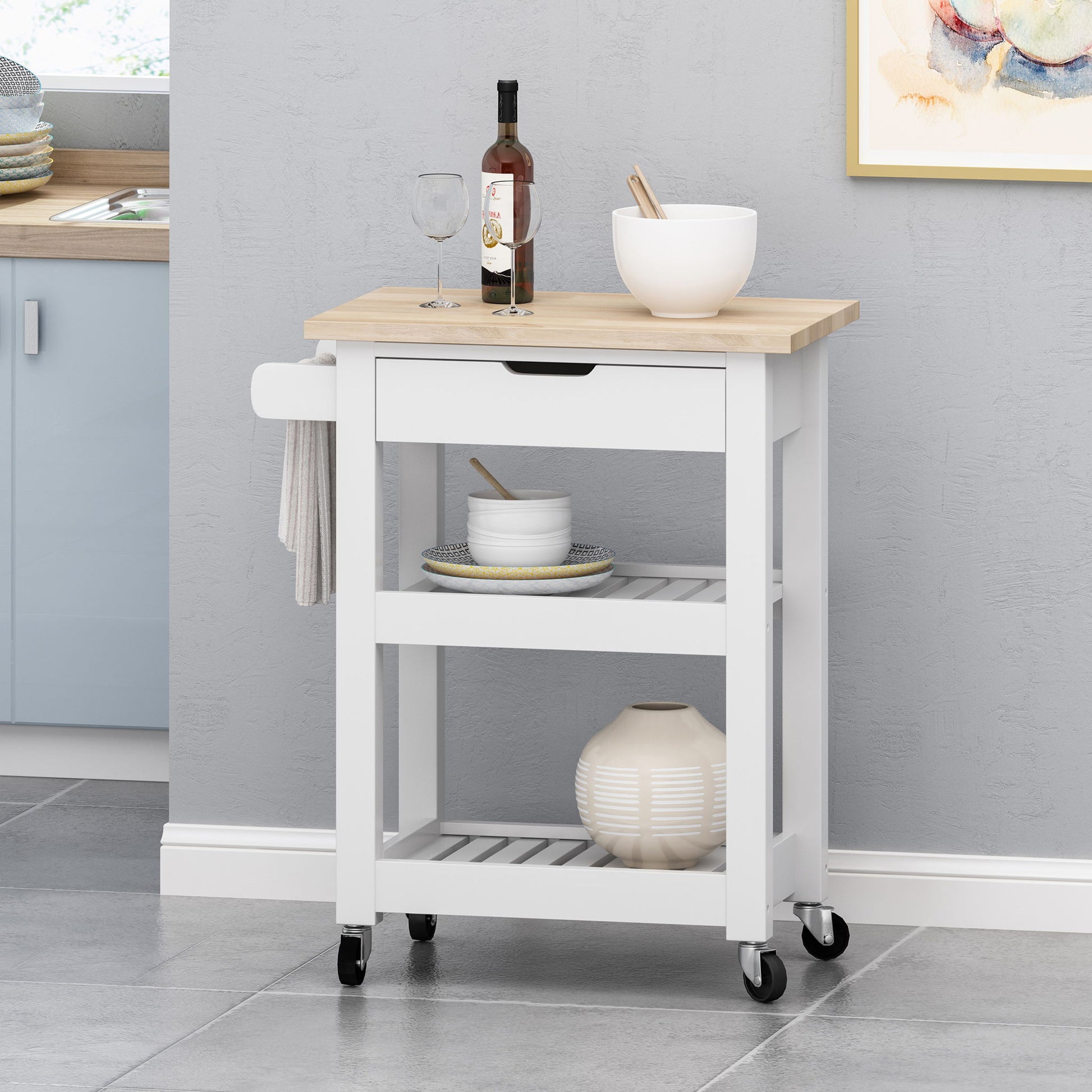 The Tattnall Kitchen Cart With Wheels features a wooden top, holds a wine bottle, glass, and white bowl, and stands against a gray wall. Its shelves store dishes and a vase, with a towel rack on the side for convenience and easy mobility.