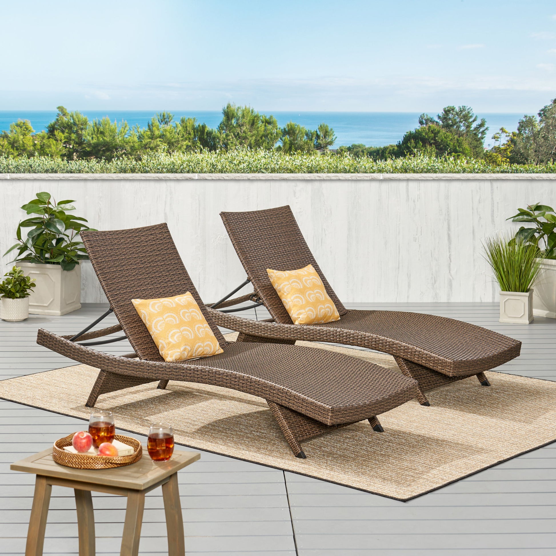 Two Salem - Chaise Lounge chairs with yellow patterned pillows sit on a beige rug on a patio. Potted plants surround the area, with a small tray of drinks and fruit nearby. Trees and an ocean view complete the relaxing scene.