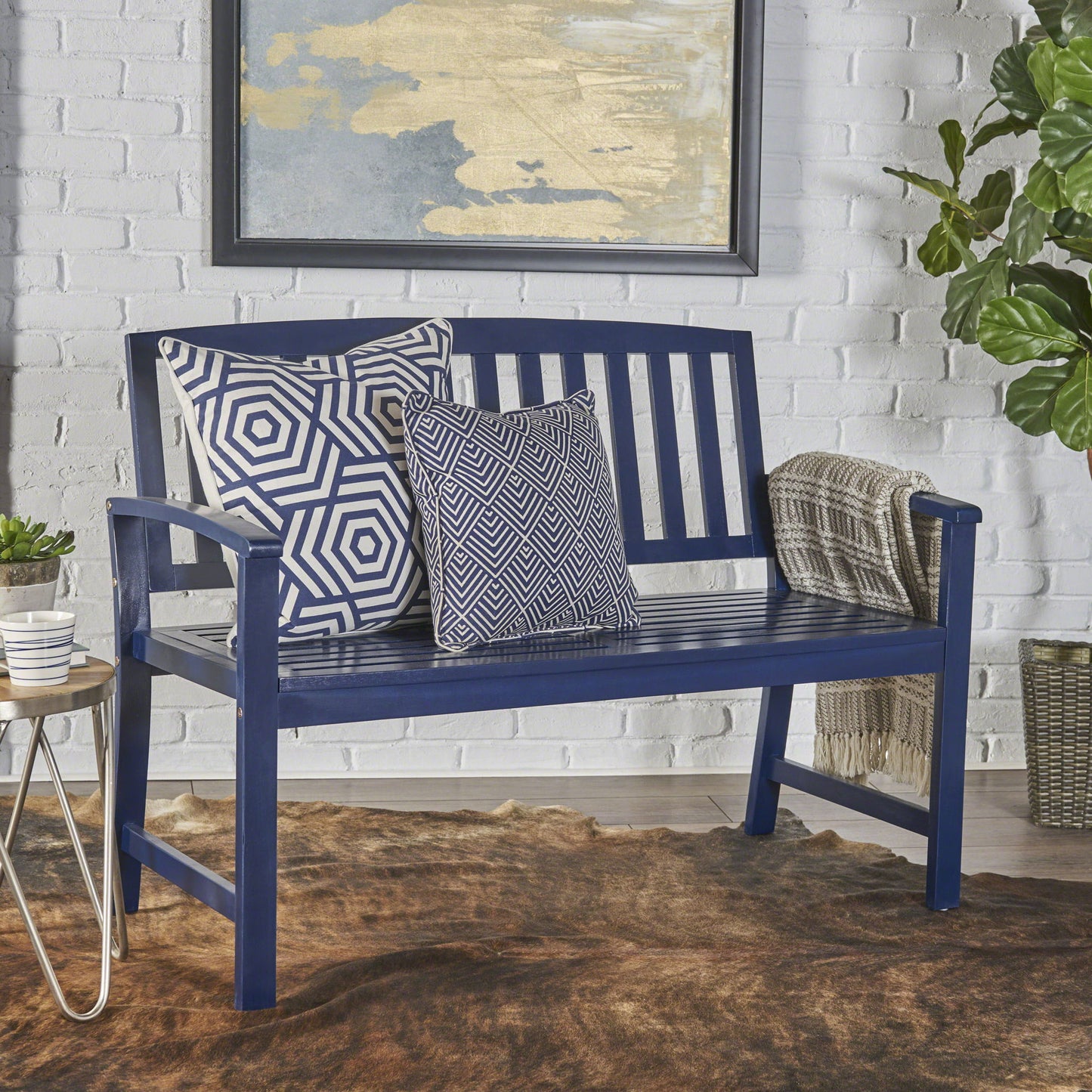The Farmhouse Style Loja Bench Indoor / Outdoor in navy blue, topped with geometric pillows and a folded throw, rests on a brown rug. A plant, small table, and abstract art accent the room's white brick wall for a stylish look.