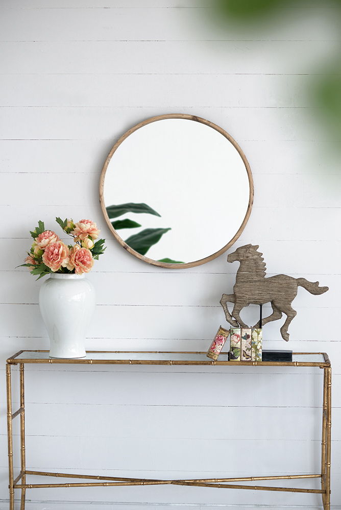 A Round Wood Mirror, Wall Mounted Mirror Home Decor For Bathroom Living Room, hangs on a white shiplap wall above a console table decorated with a vase of pink flowers, books, and a horse figurine. Green leaves are reflected in the mirror.