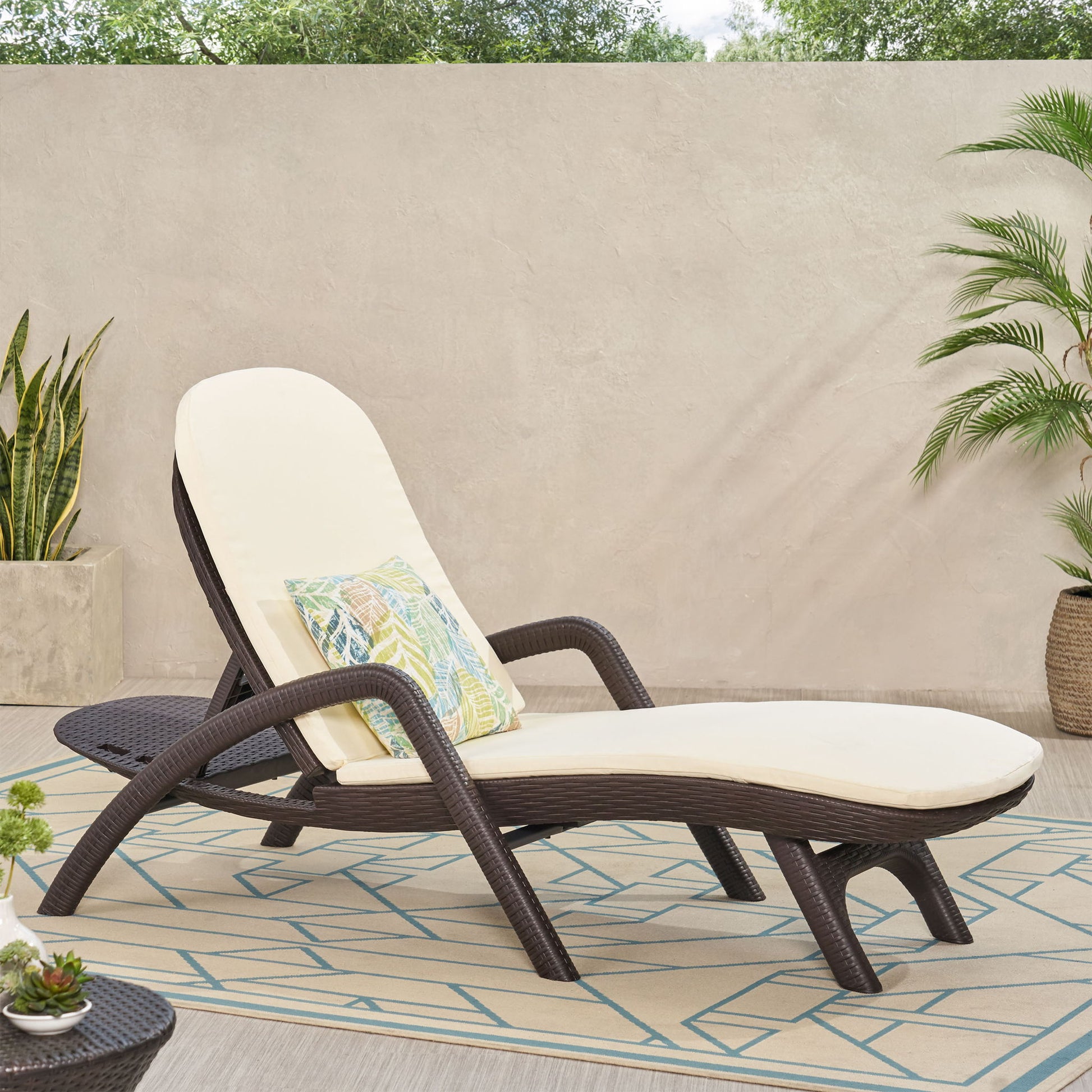 The Mikael - Chaise Lounge With Cushion, featuring brown wicker, cream cushions, and a decorative pillow, rests on a patterned outdoor rug with potted plants by a beige wall in a sunny patio setting.