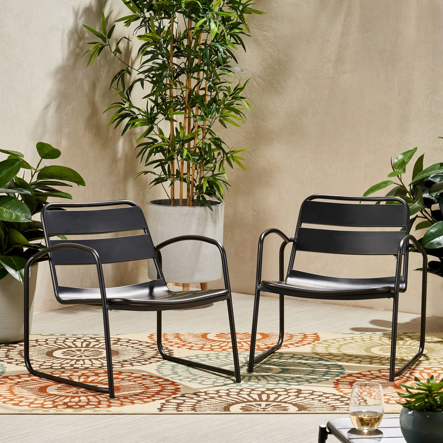 Two Conrad Outdoor Chairs in modern black metal are set on a vibrant patterned rug outside, surrounded by potted green plants, with a drink placed on a small corner table.
