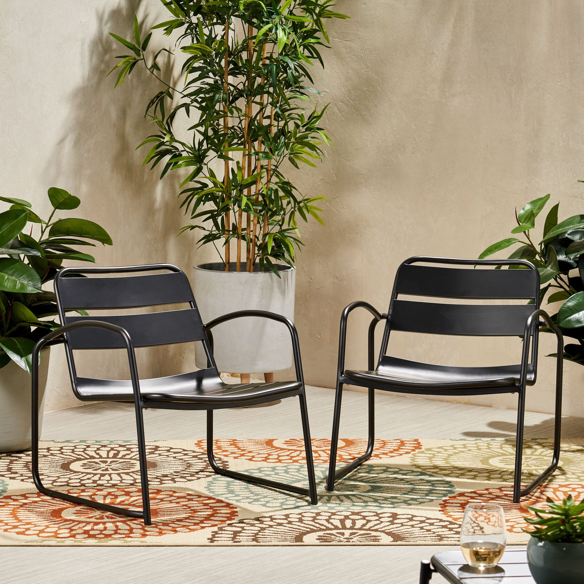 Two Conrad Outdoor Chairs in modern black metal are set on a vibrant patterned rug outside, surrounded by potted green plants, with a drink placed on a small corner table.