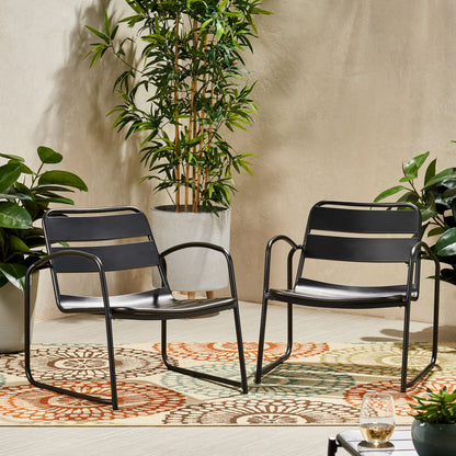 Two Conrad Outdoor Chairs in modern black metal are set on a vibrant patterned rug outside, surrounded by potted green plants, with a drink placed on a small corner table.