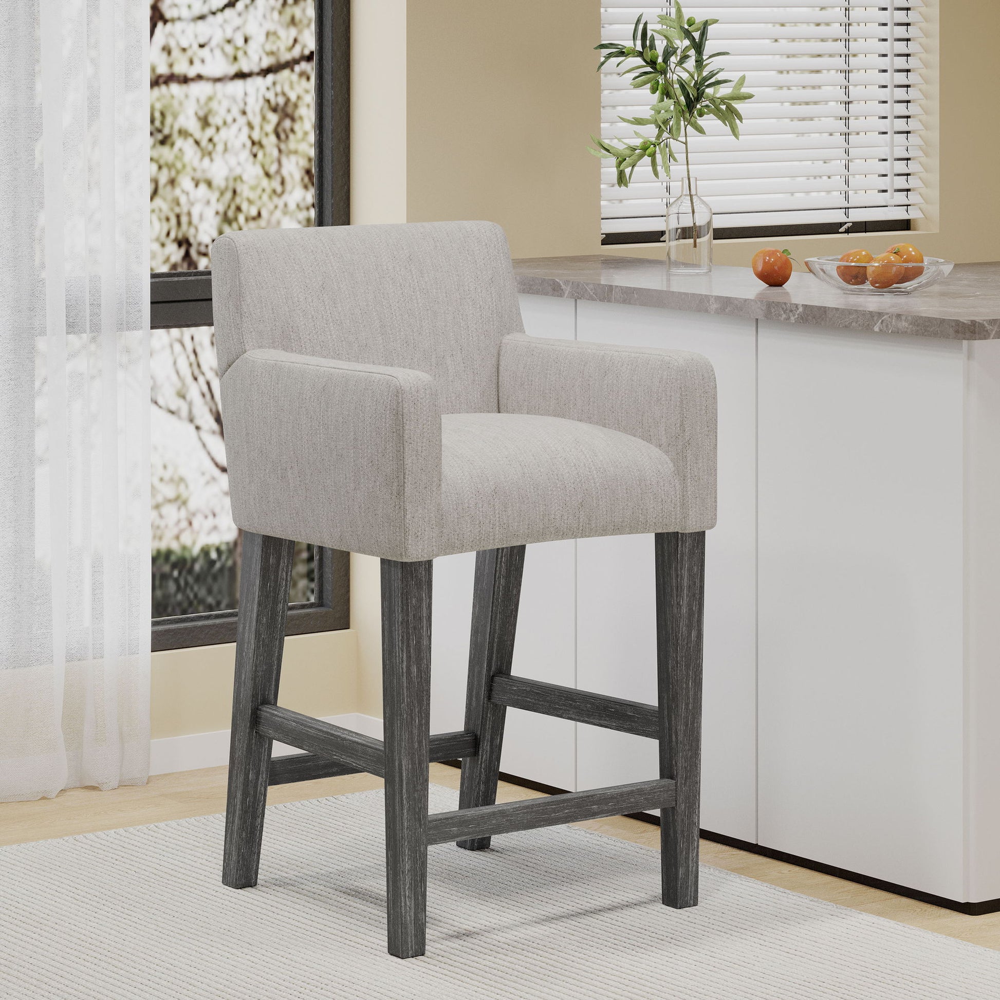 The Charmed Upholstered Counter Stool With H Frame Base And Round Arms, featuring light gray fabric and dark wooden legs, sits in a bright kitchen near a window beside a counter with a plant and oranges.